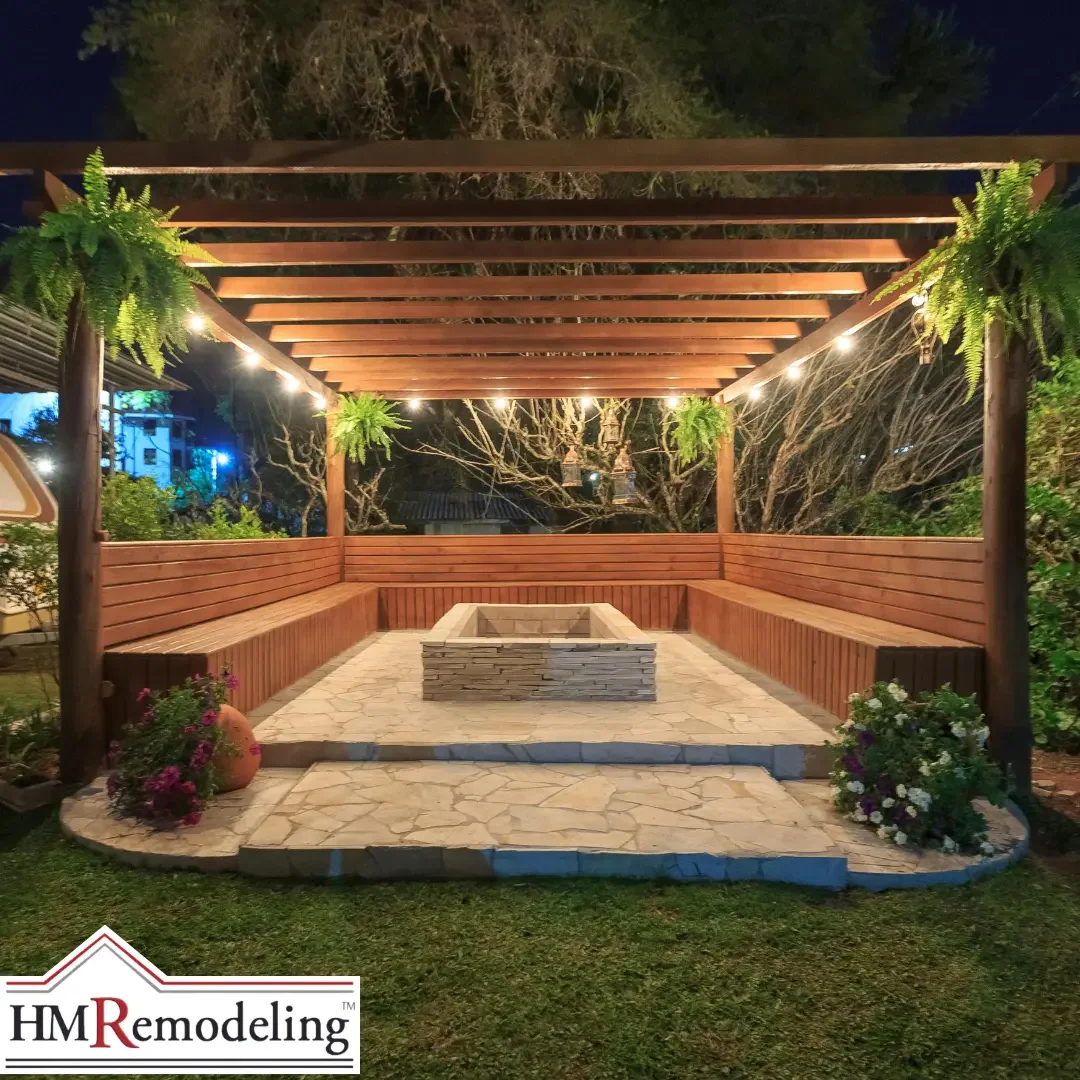 A wood pergola lit with string lights at night, sheltering a stone fire pit flanked by built-in benches and flowers, an outdoor entertainment area concept for a Michiana area home.