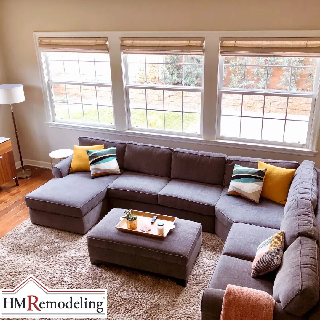 A bright South Bend, Indiana living room with freshly painted warm beige walls, white wood trim, three large double-hung windows with Roman shades, hardwood floors, and a large gray sectional sofa with colorful accent pillows