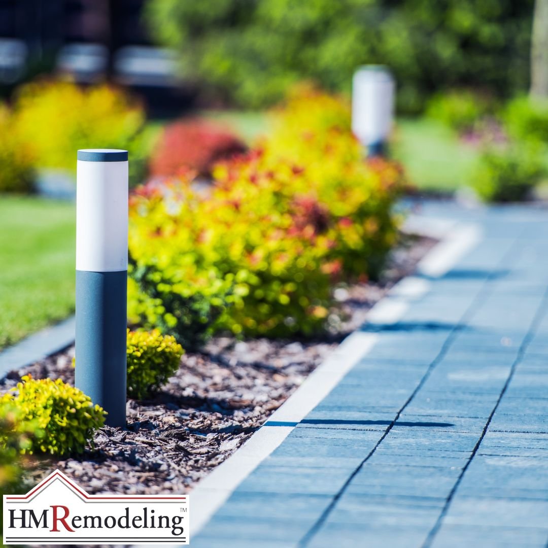 Modern dark gray bollard path light along a stone paver walkway bordered by a mulched landscape bed with colorful shrubs, exterior lighting upgrade serving South Bend and Northern Indiana
