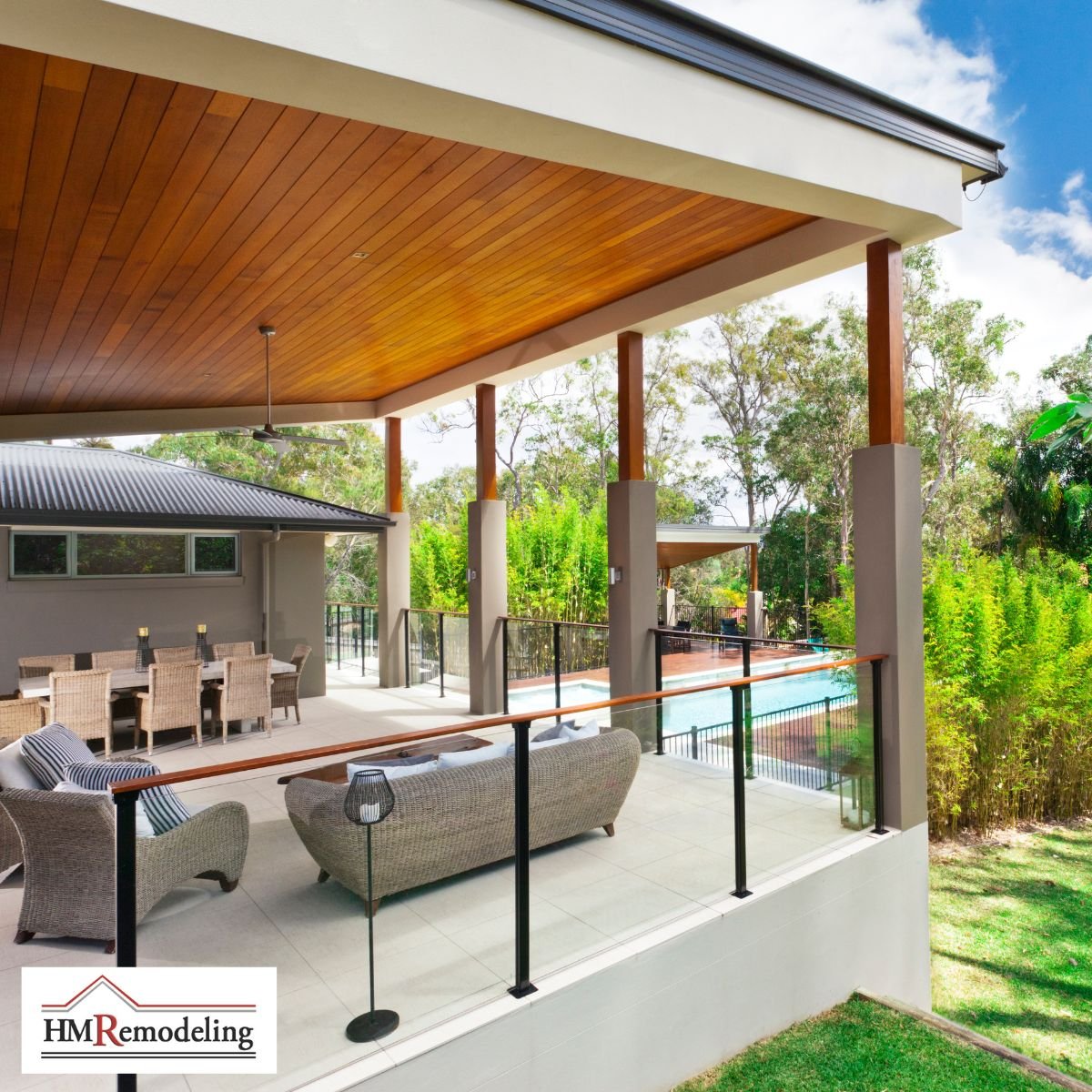 Modern covered outdoor patio with wood plank ceiling, concrete columns, glass panel and wood cap railing, wicker lounge seating, outdoor dining area, and backyard pool view by HM Remodeling in South Bend, Indiana