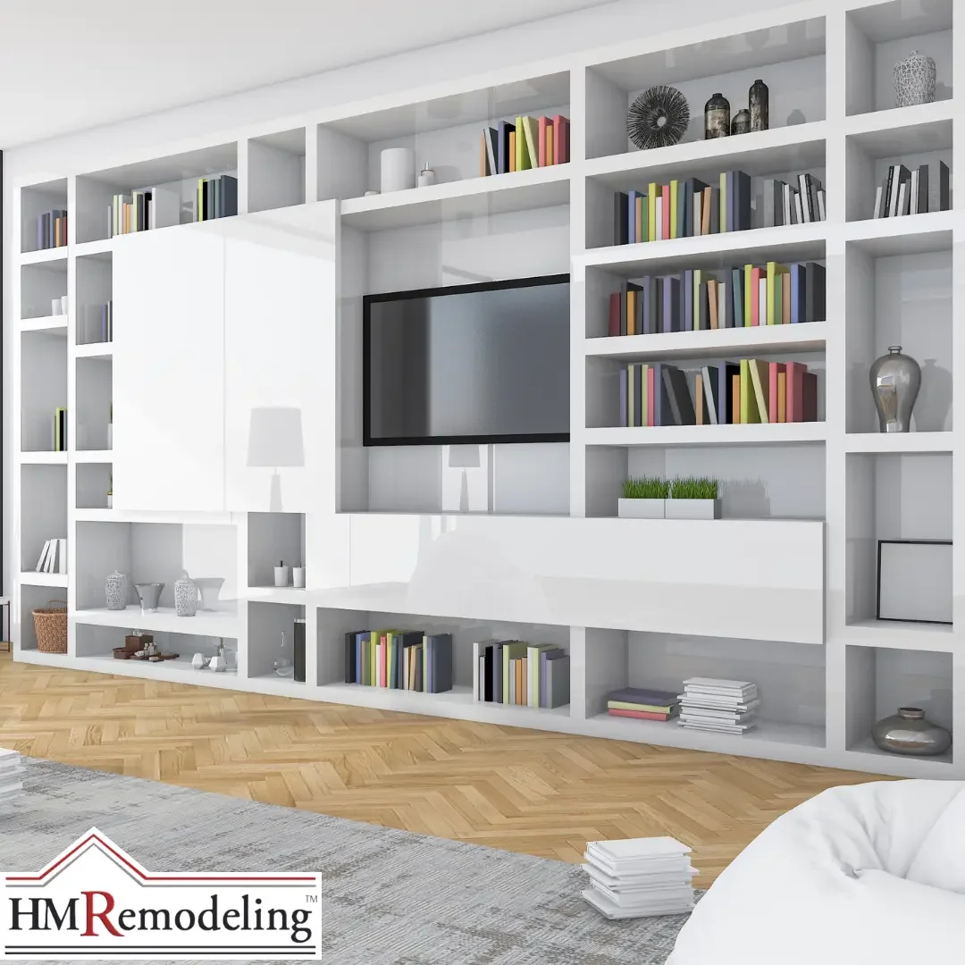 A modern floor-to-ceiling white built-in wall unit with open shelving filled with colorful books and decor and a centrally mounted flat-screen television, set above herringbone hardwood flooring in a South Bend, Indiana living room