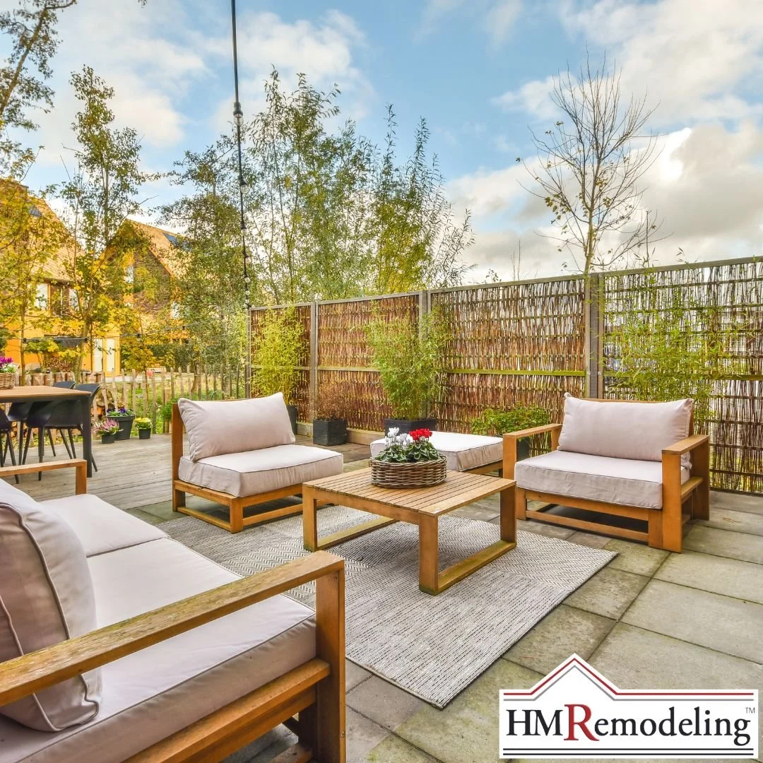 Composite deck with gray wicker sectional sofa, upholstered ottoman, round boho area rug, and potted pink flowers set against a wooded backyard in South Bend, Indiana