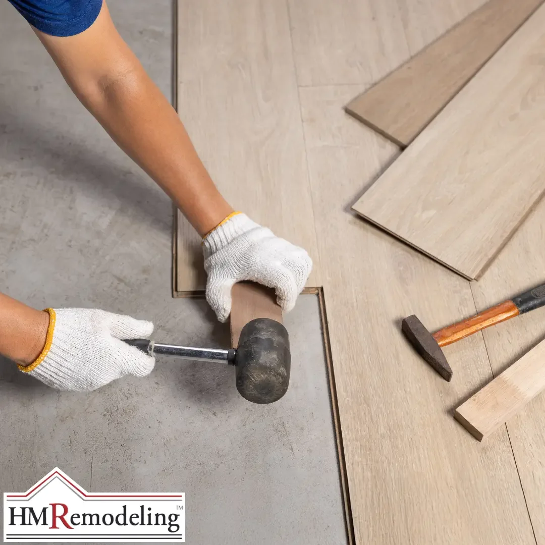A gloved installer using a rubber mallet and tapping block to fit light oak hardwood flooring planks together on a concrete subfloor in a South Bend, Indiana home, with additional planks and a hammer visible nearby