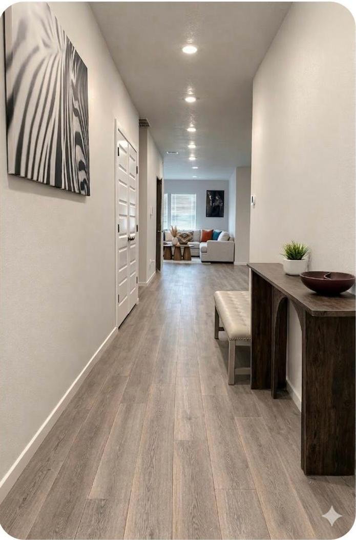 Interior hallway leading to a living room with white sofa, black side tables, and wall art, with wooden flooring, white walls, and ceiling lights.