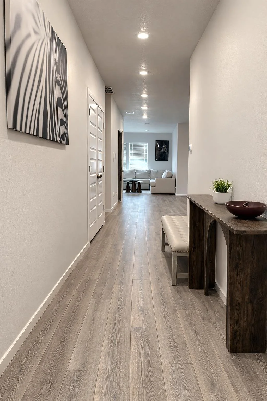 Interior hallway leading to a living room with white sofa, black side tables, and wall art, with wooden flooring, white walls, and ceiling lights.
