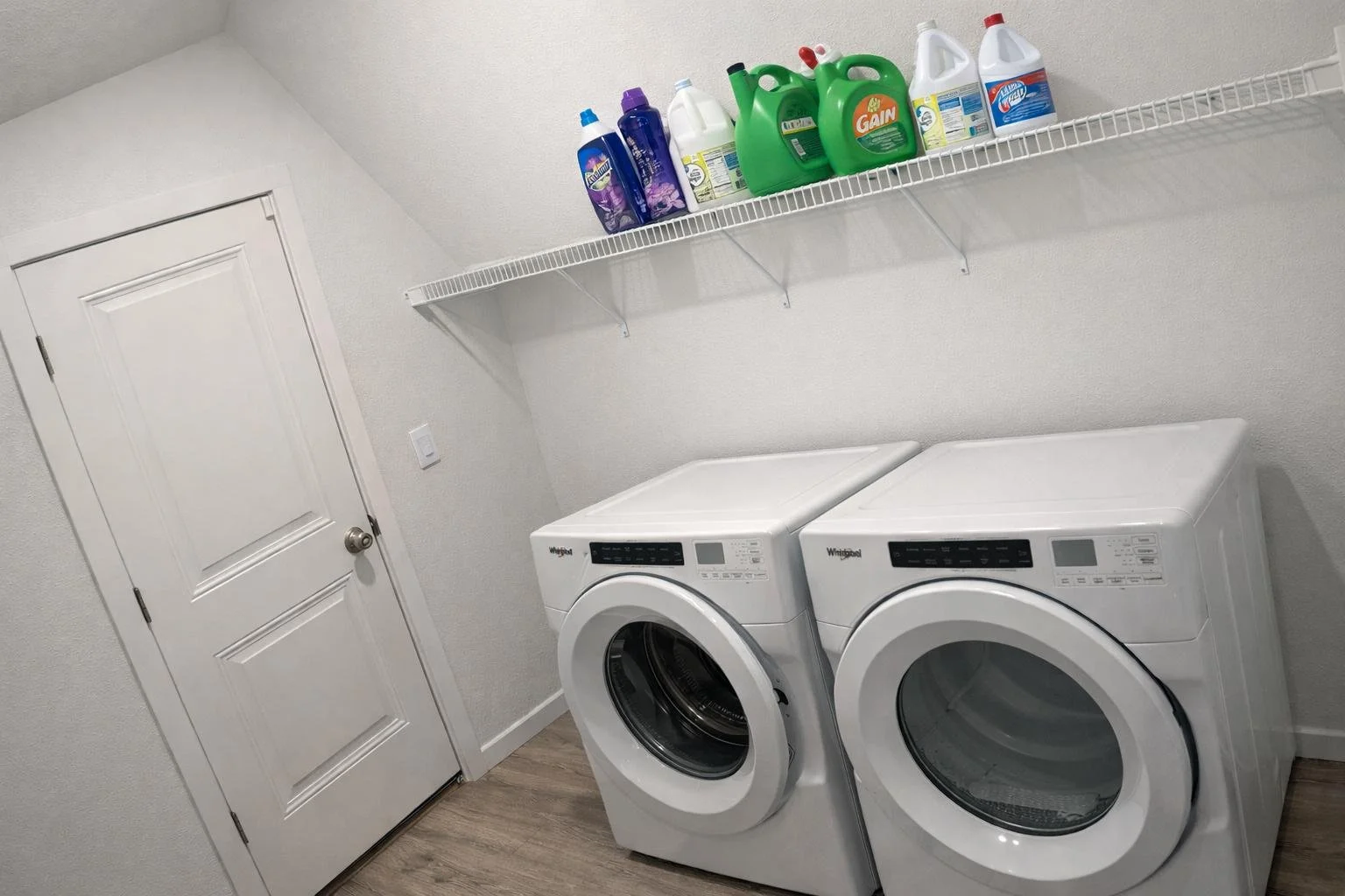 Laundry room with a white washing machine and dryer side by side, a white wire shelf above holding detergent bottles, and a closed door on the left wall.