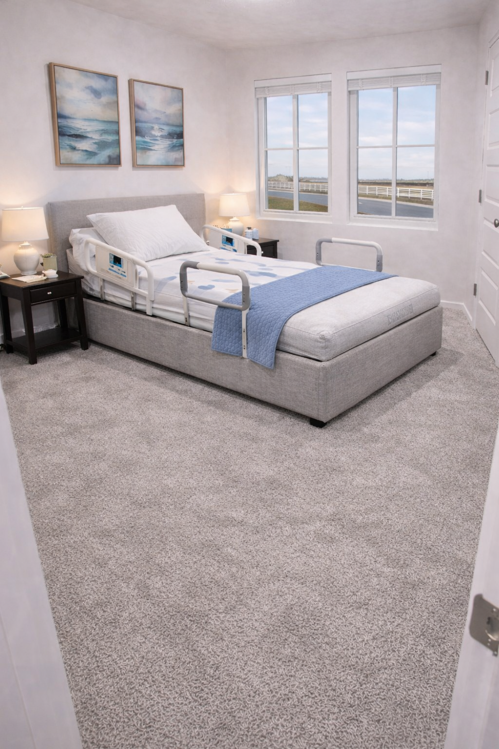 A hospital room with a bed, bedside tables with lamps, and windows showing an outdoor scene with a road and fencing.