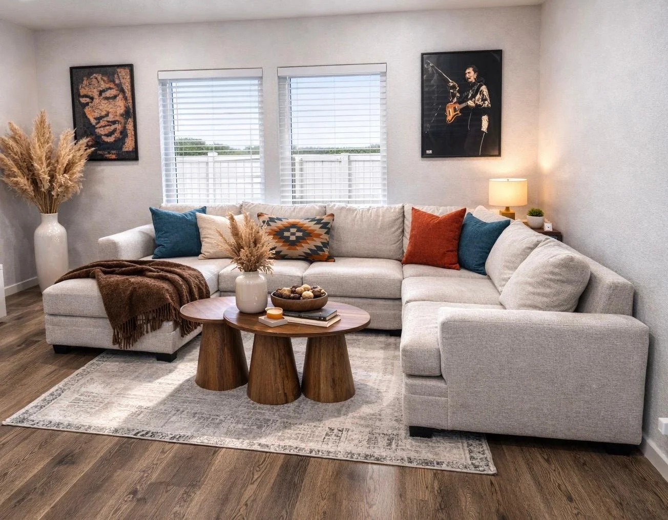 Living room with a large sectional sofa, a flat-screen TV on a white stand, two round wooden tables with a candle and tray, framed art on white walls, and windows showing an outdoor scene with red buildings and trees.