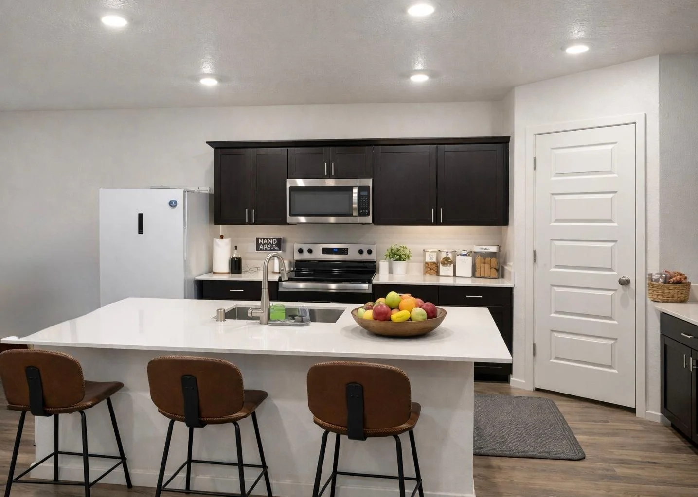 Modern kitchen with white island counter, brown leather stools, black cabinets, stainless steel microwave and stove, white refrigerator, bowl of fruit, and various kitchen supplies.