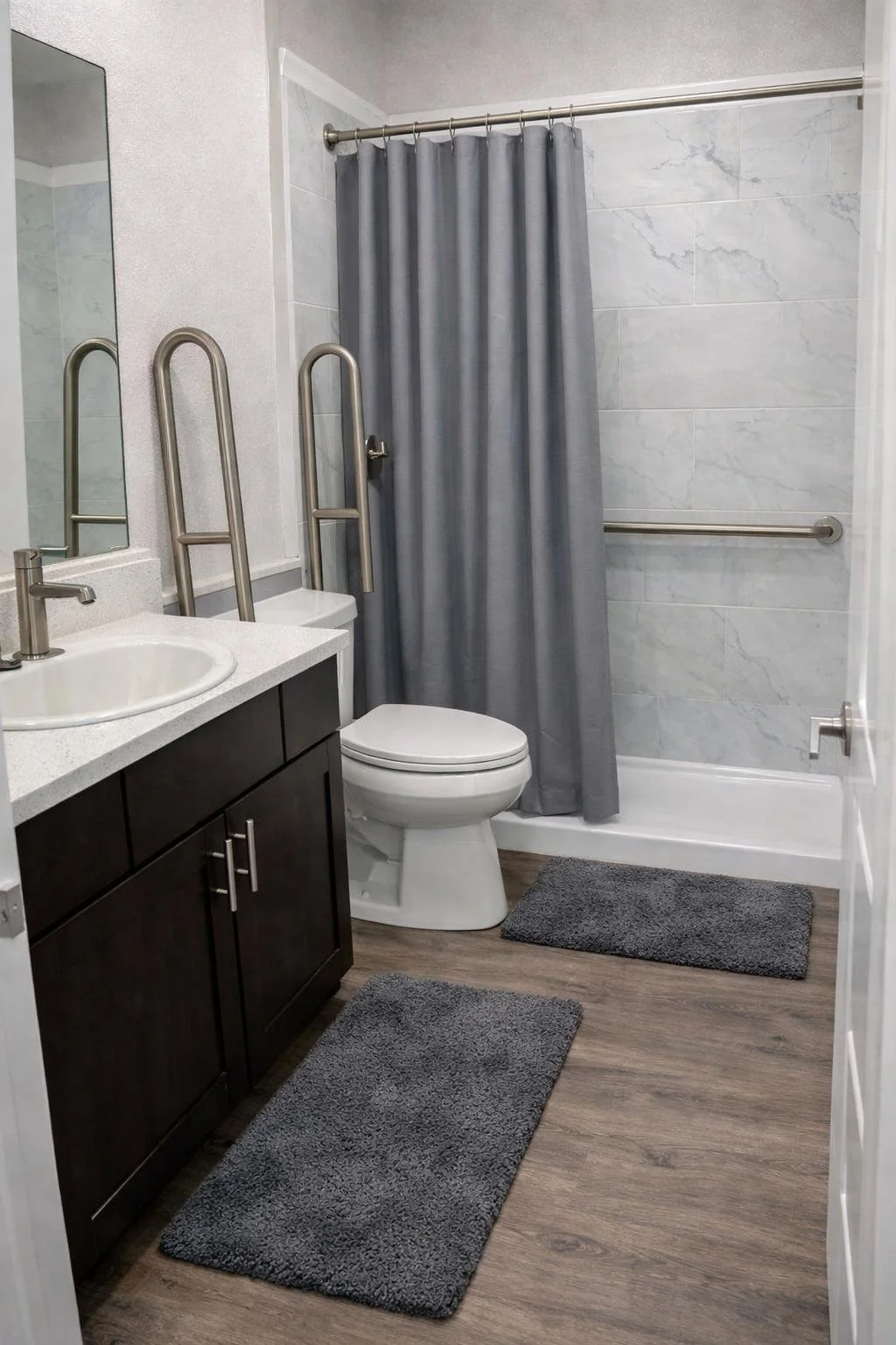 Accessible bathroom with grab bars, gray shower curtain, dark wood vanity, white sink, gray textured rugs, and wood-like flooring.