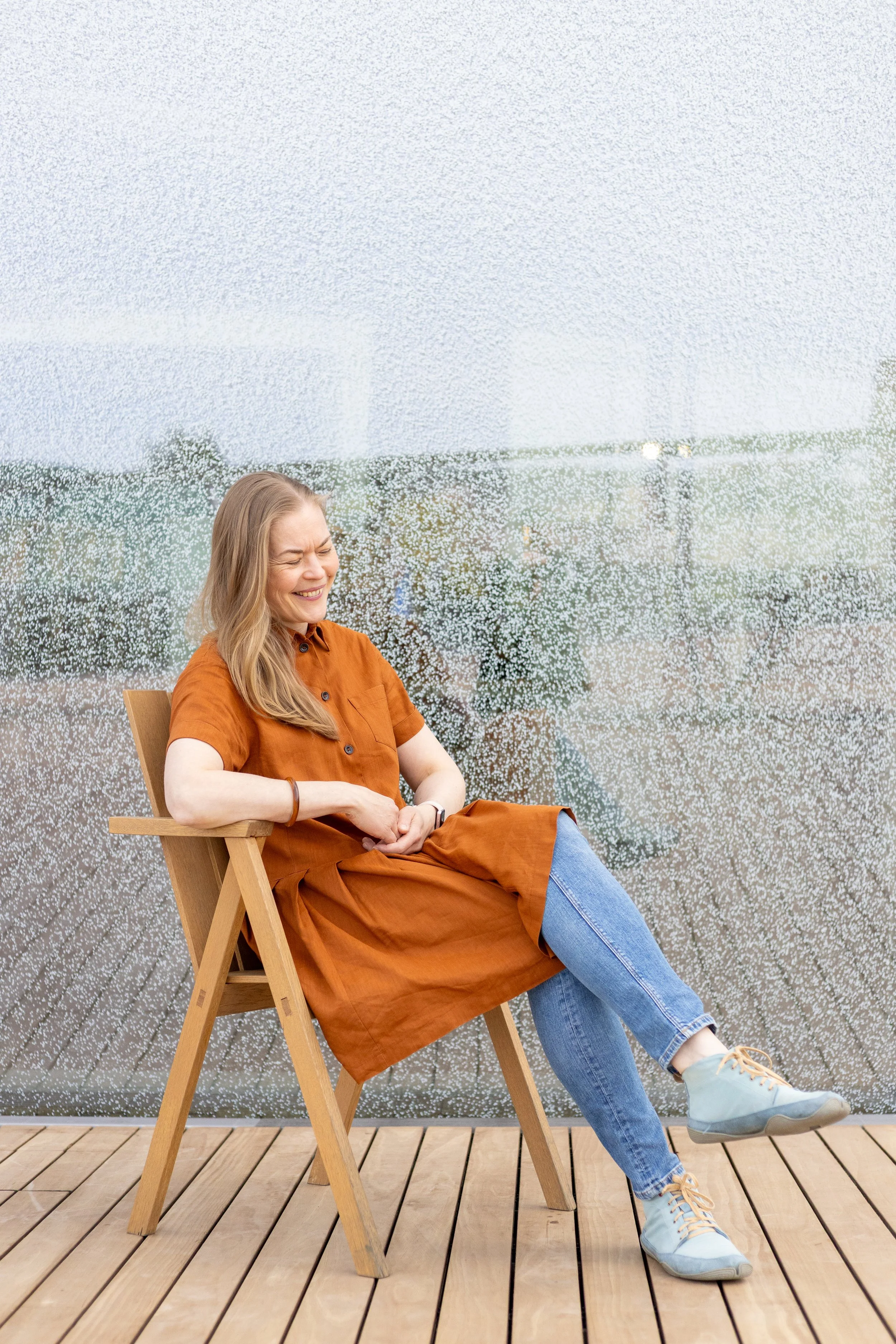 Pauliina Ilonoja is sitting on a wooden chair with a happy expression, wearing a rust-colored dress, blue jeans, and light blue sneakers, sitting on a wooden deck with a textured glass wall in the background. Picture: Kristiina Kaitala