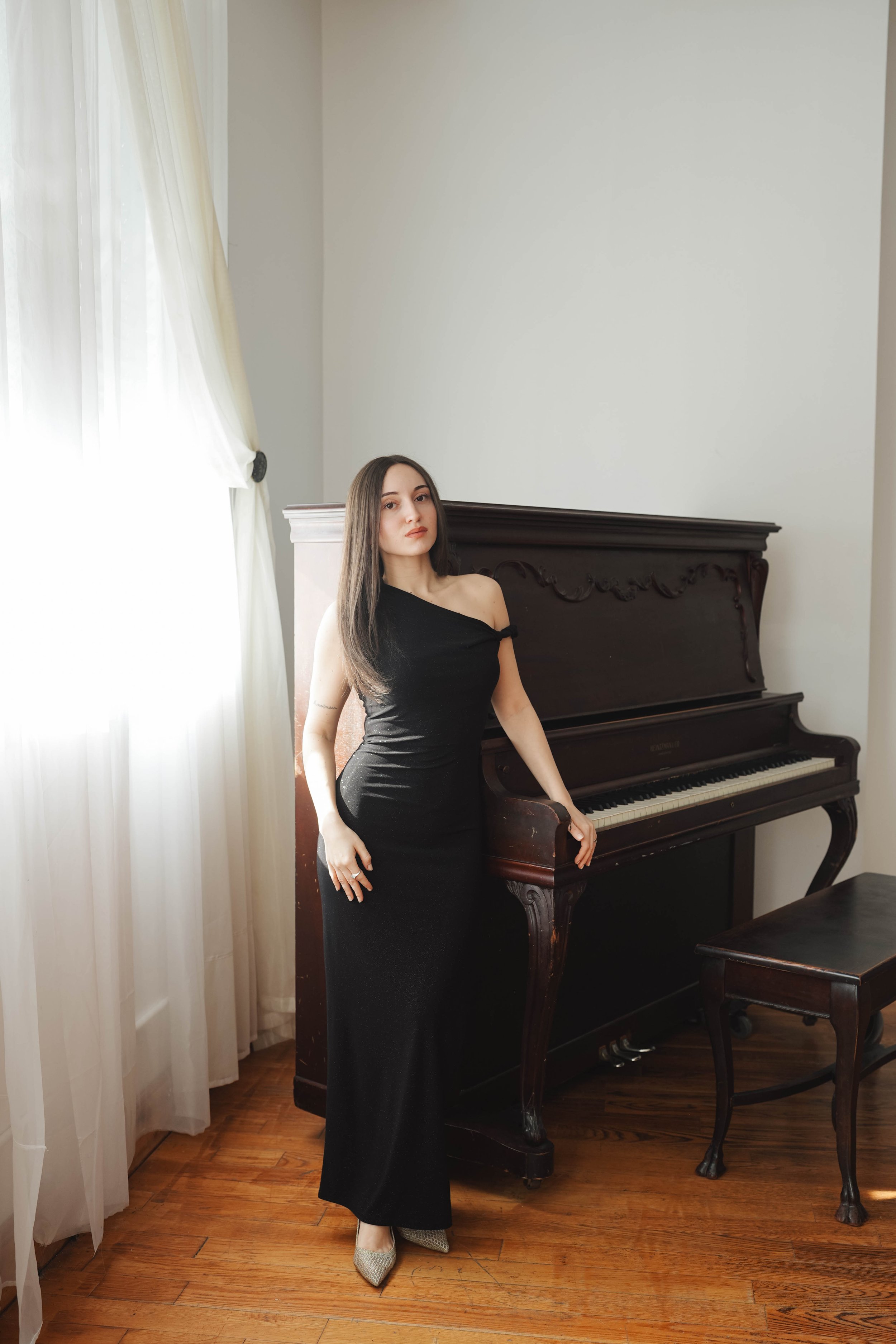 A woman in a black off-shoulder dress standing next to an antique upright piano in a room with hardwood floors and white walls. Light coming through sheer curtains on a window.