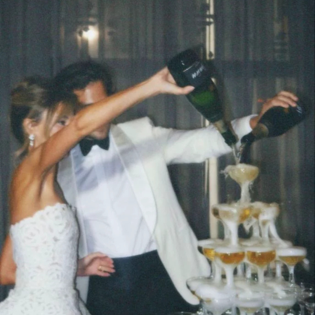 A bride and groom pouring champagne into a pyramid of glasses at a wedding celebration.