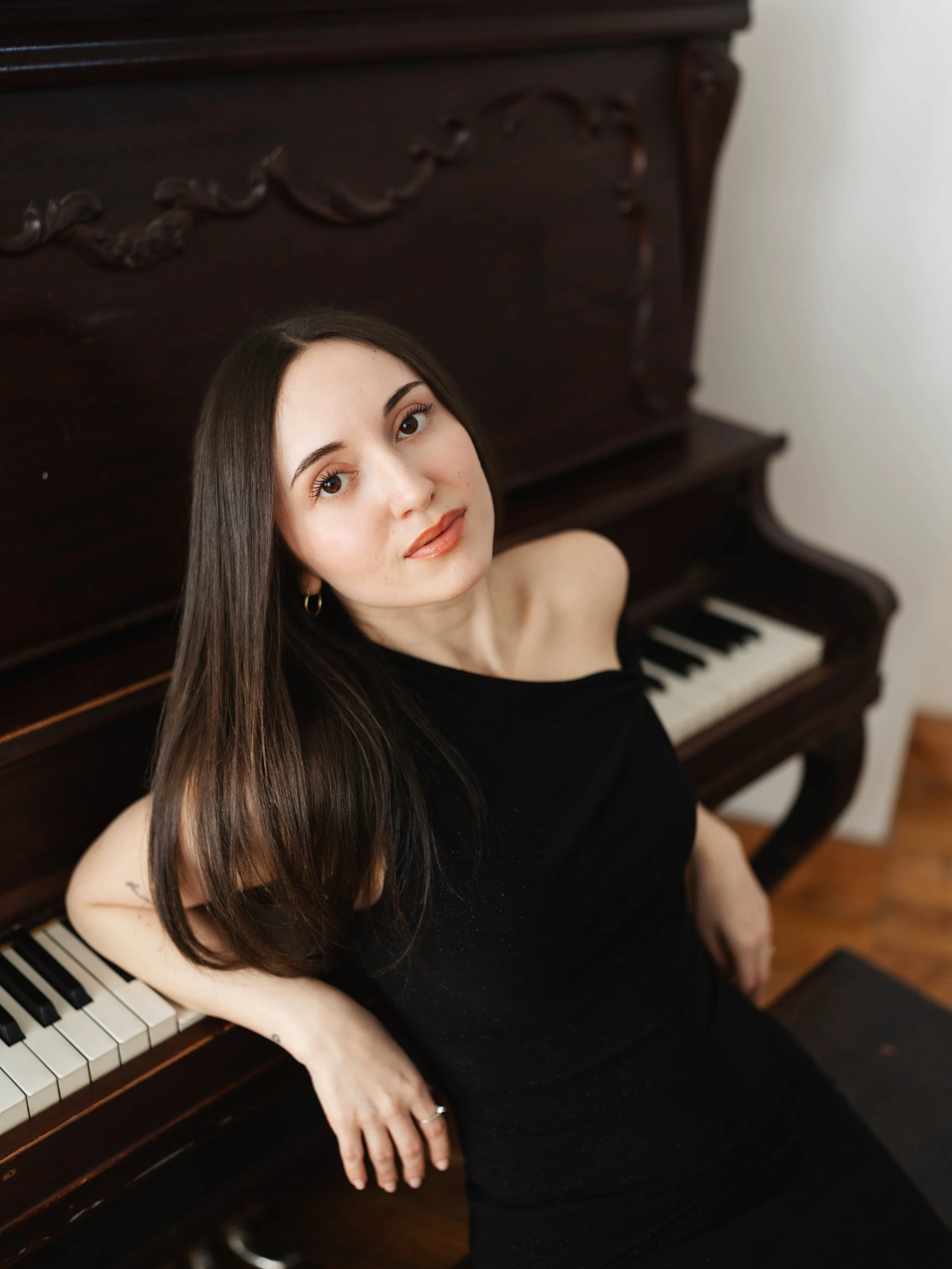 A young woman with long dark hair, wearing a black dress, sitting at an upright piano with dark wood finish, looking up at the camera with a relaxed expression.