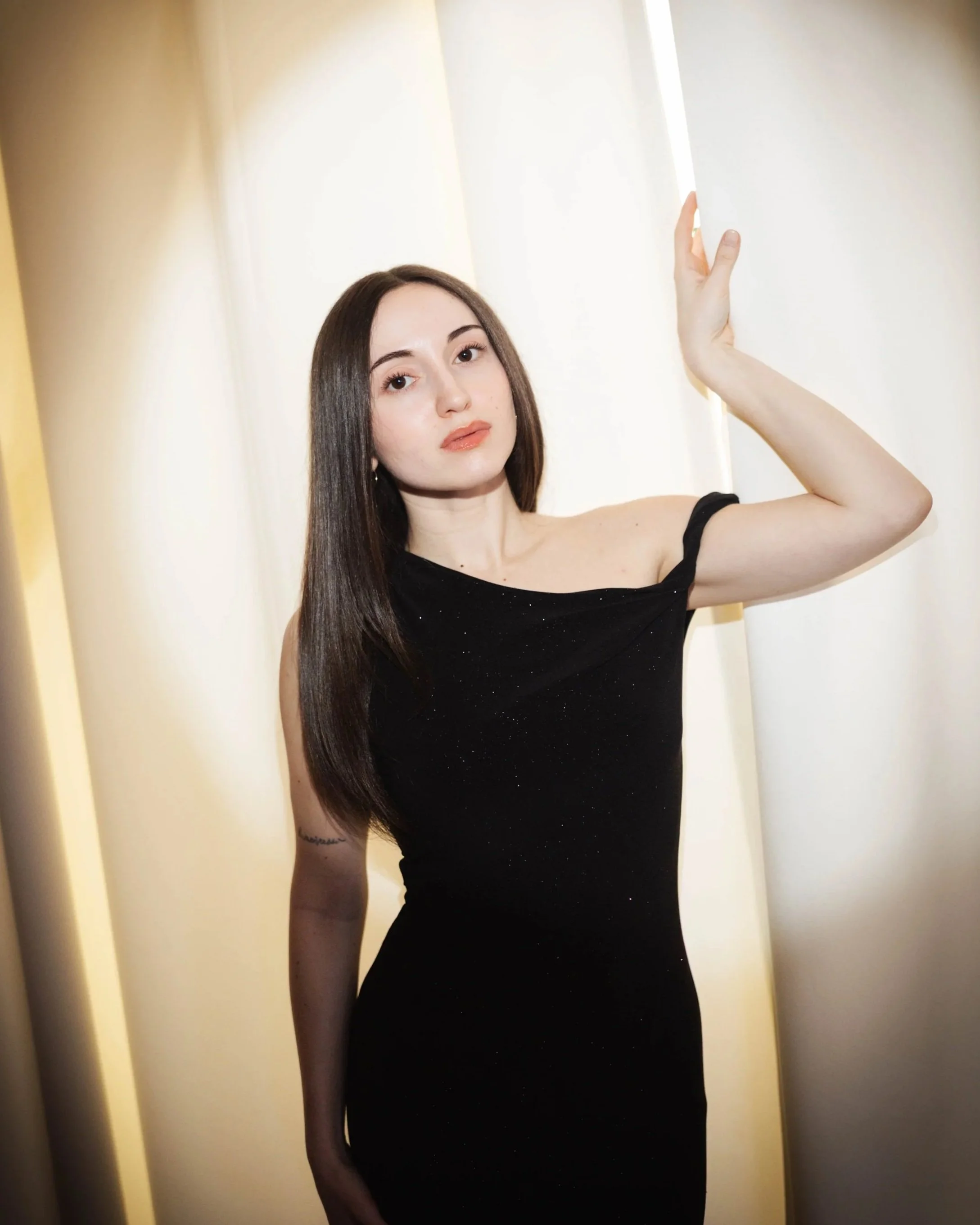 A young woman with long dark hair, wearing a black off-shoulder dress, standing in front of a light-colored background, touching a wall or curtain with her right hand.
