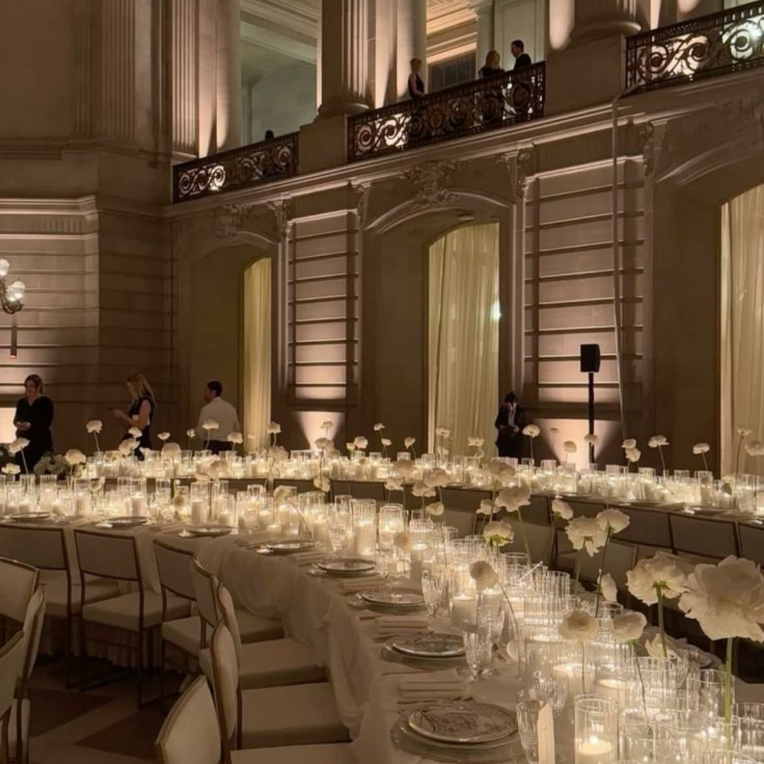 Elegant banquet hall with long table set with white flowers, candles, glassware, and plates; high ceilings with ornate architectural details, tall columns, and a balcony with people standing and talking.
