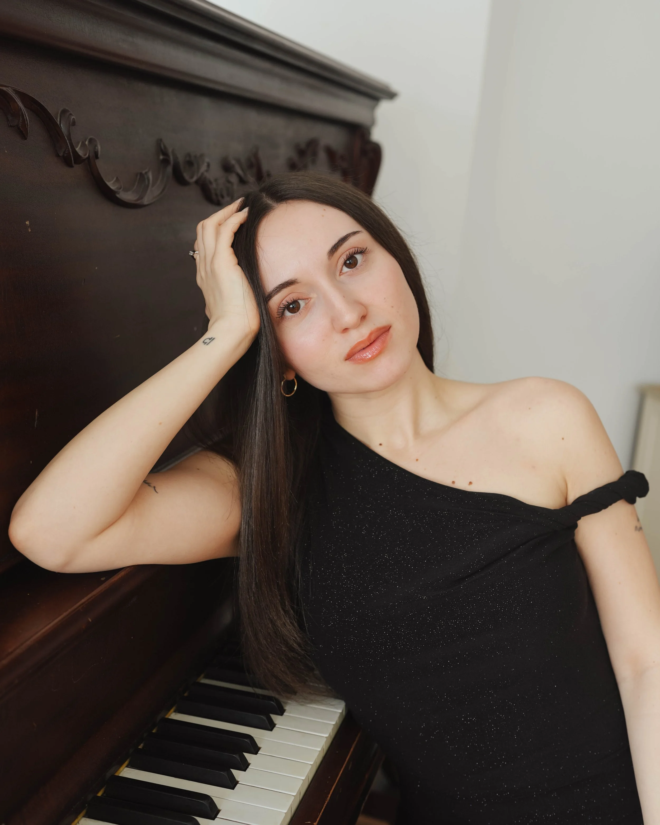 A woman with long dark hair and light skin, wearing a black off-shoulder top with sparkles, leaning against a wooden piano with her hand on her head, looking at the camera.