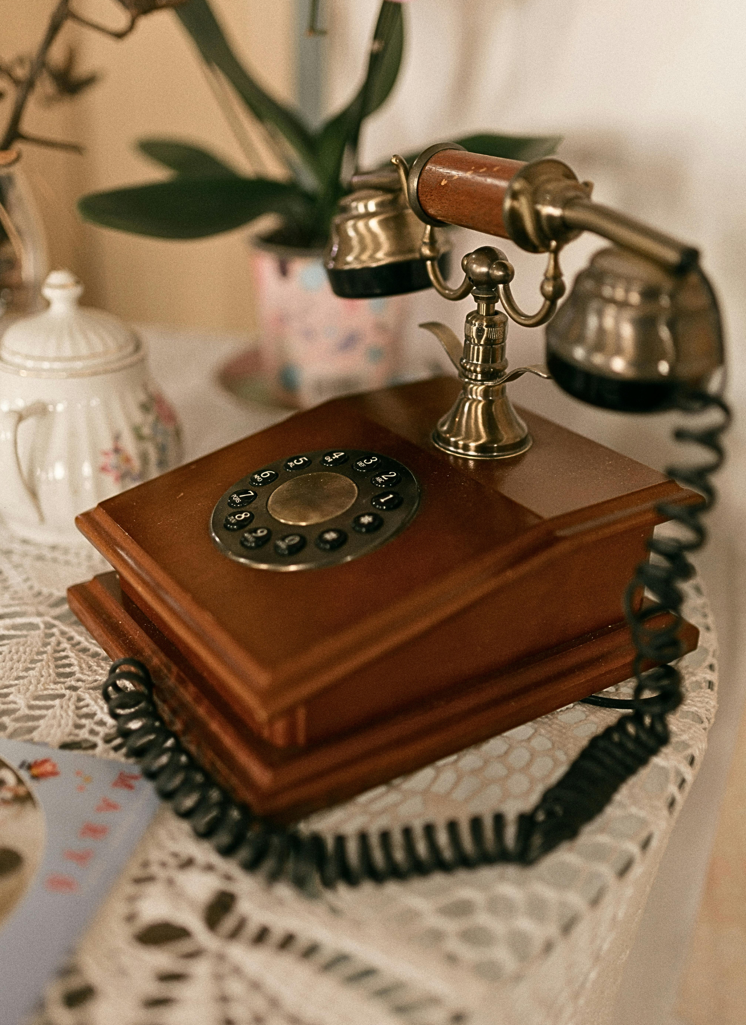 Vintage rotary dial telephone on a wooden base, placed on a lace tablecloth with a blurred decorative jug and potted plant in the background.