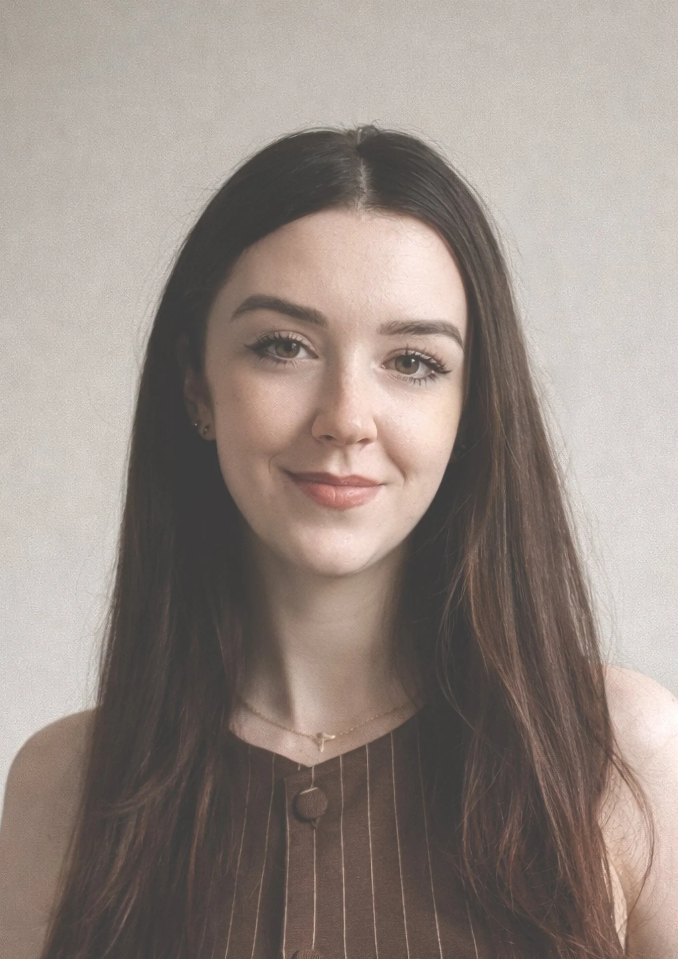 A young woman with long brown hair, wearing a sleeveless brown top with thin white pinstripes and a small pendant necklace, smiling at the camera against a neutral background.
