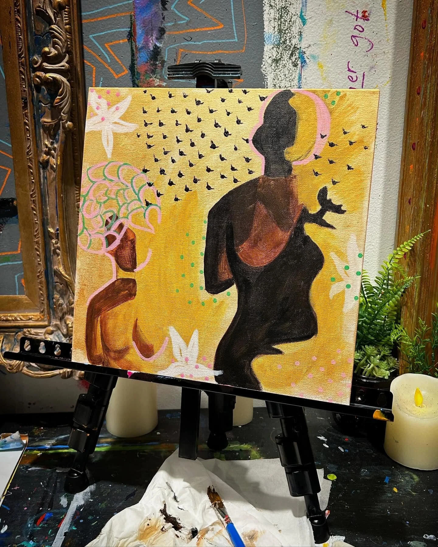A colorful painting of a woman with short hair, sitting with her back slightly turned, surrounded by abstract shapes, small black birds, and white butterflies on a yellow background. The painting is set on an easel in an artist's studio.