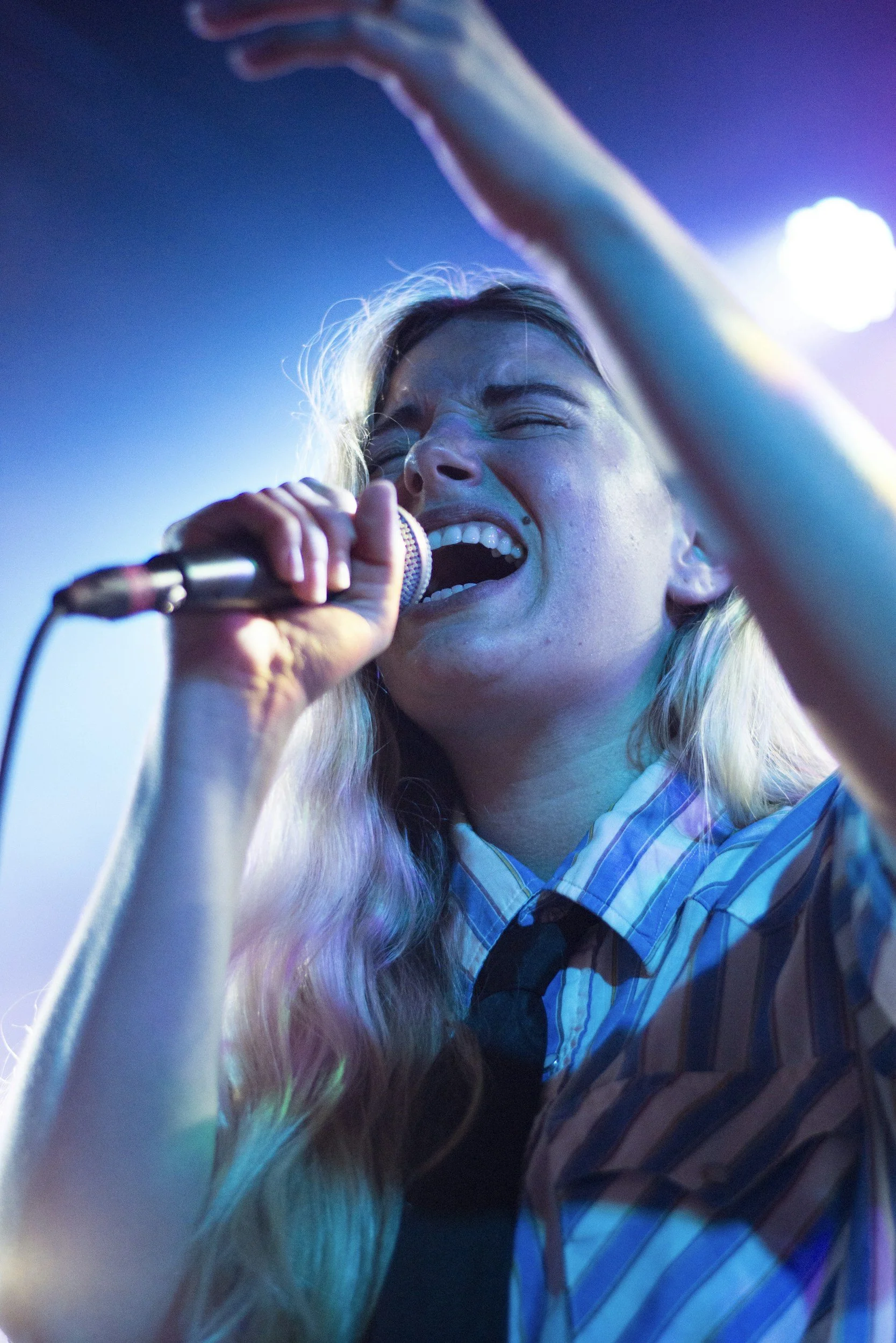 A young woman with long blonde hair passionately singing into a microphone at a concert, wearing a striped shirt, with colorful stage lighting illuminating her face.