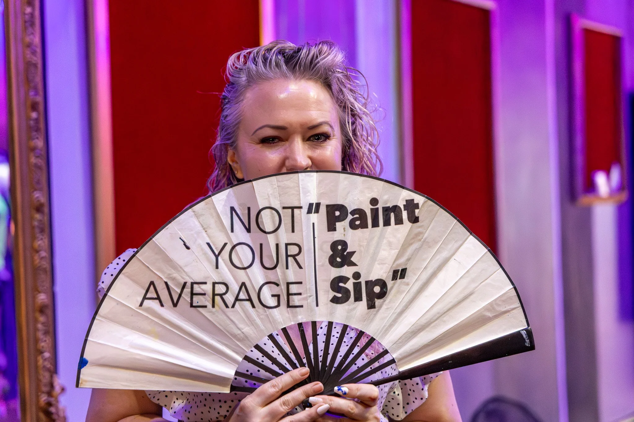 A woman with blonde, curly hair mockingly hiding behind a fan with humorous writing, in a colorful indoor setting.