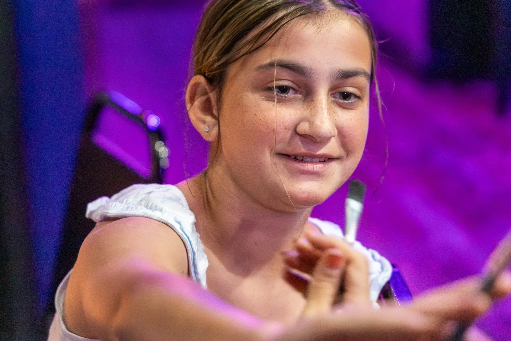 A young girl with blonde hair and freckles smiling, with a purple background, wearing a white sleeveless top.