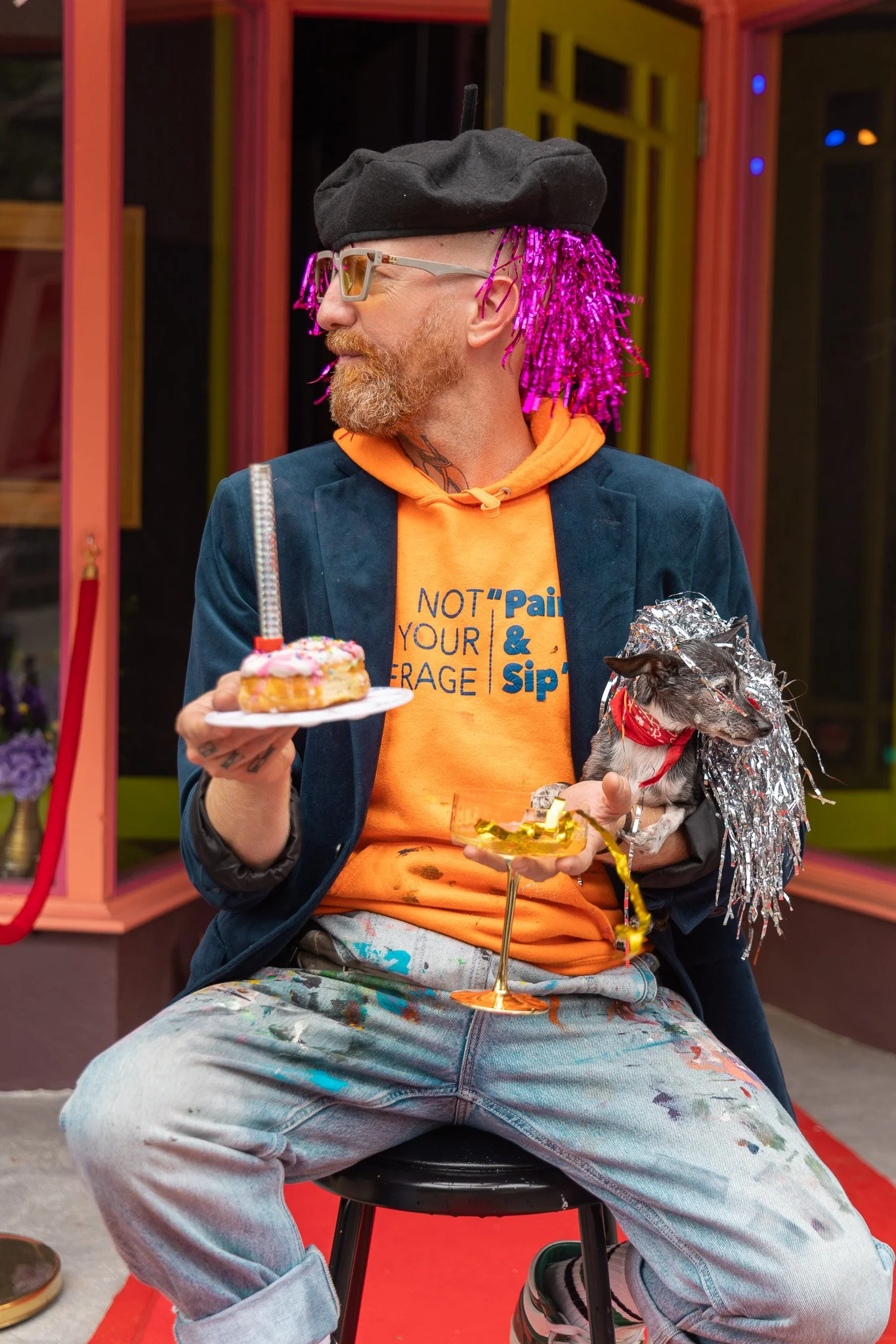 A man with a beard, bright pink hair extensions, wearing sunglasses, a black beret, an orange hoodie, a dark jacket, and baggy paint-splattered jeans, sitting on a stool holding a cupcake and a drink, with a small dog wrapped in silver tinsel in his lap, in front of colorful background.
