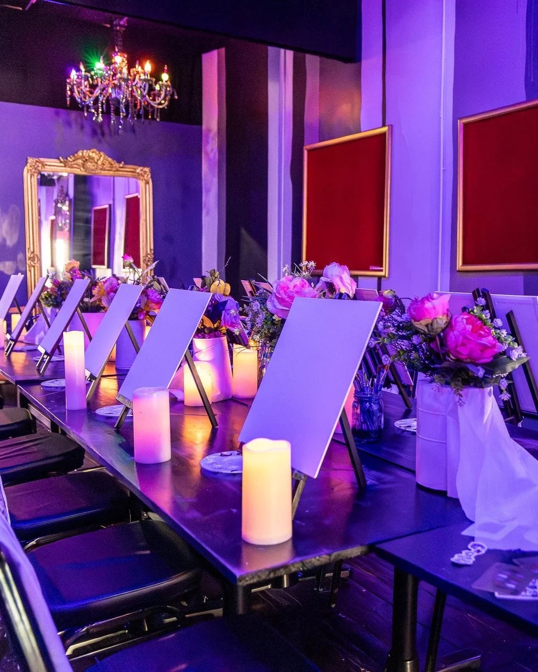 A decorated conference room with a long table, flower arrangements, lit candles, and portrait displays on the walls, illuminated with purple and pink lighting, a chandelier hanging from the ceiling.