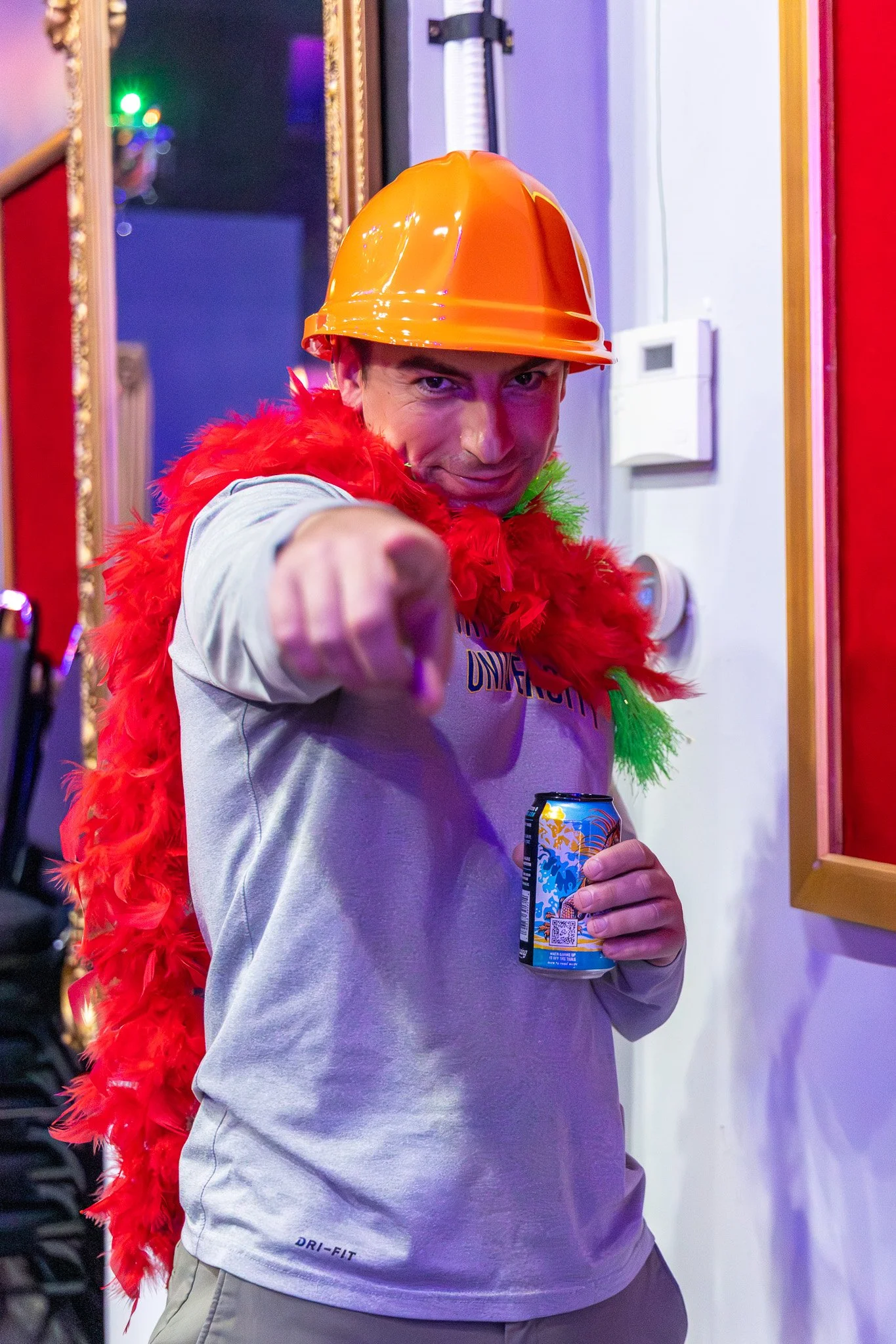 Man wearing an orange construction helmet and a red feather boa, holding a can of drink, pointing towards the camera, indoors with colorful lighting.