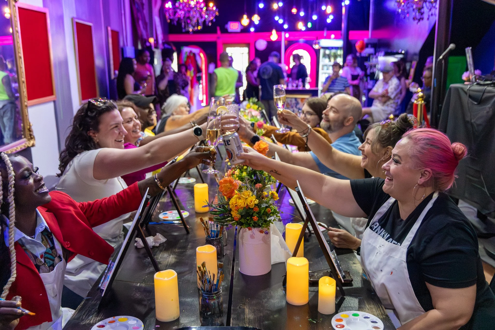 People sitting at a long table, raising glasses in a toast, with colorful decorations and candles, in a lively indoor party setting.
