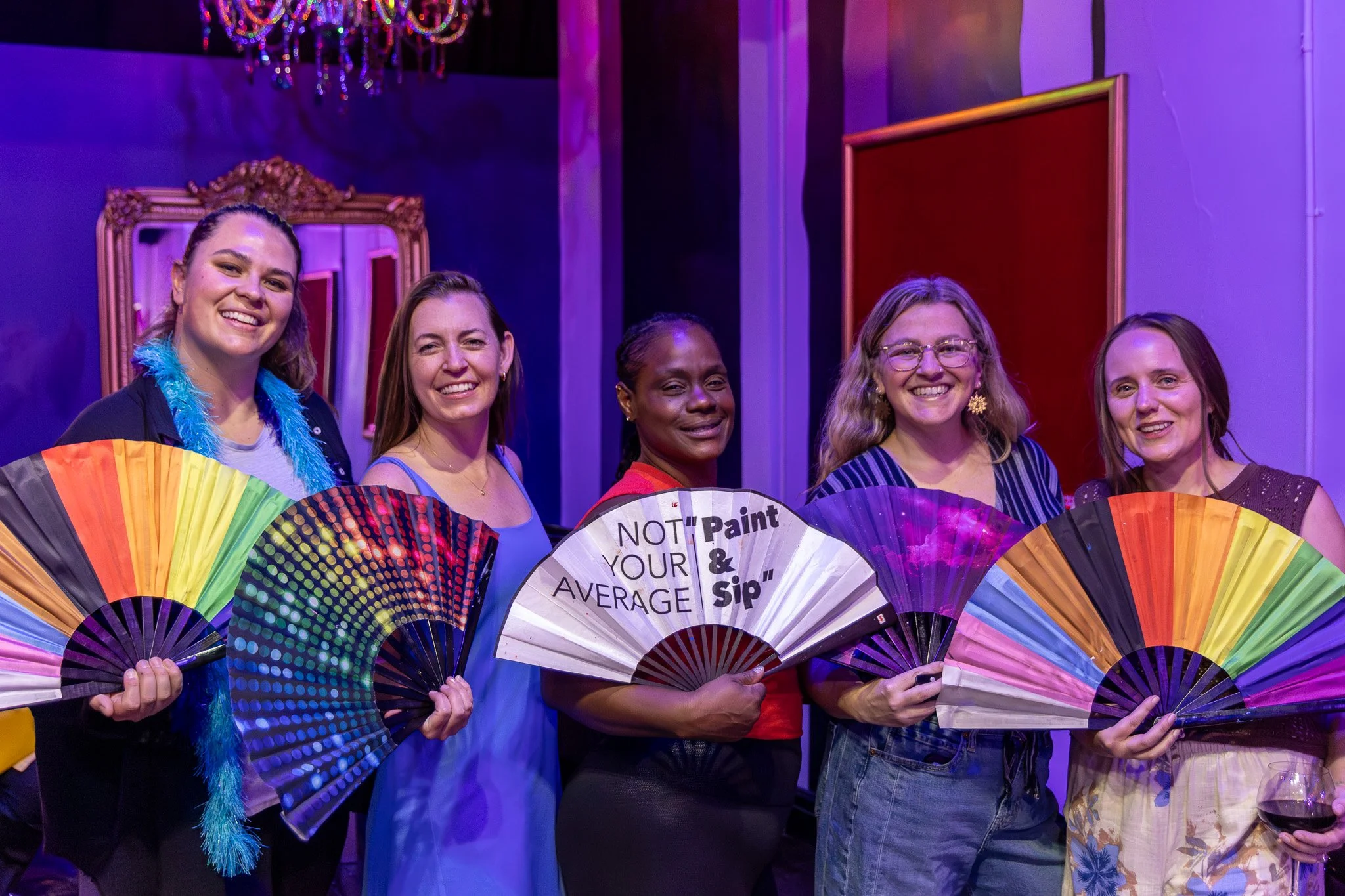 A group of five women standing together, holding rainbow-colored fans, smiling, with a chandelier overhead and purple lighting in the background.