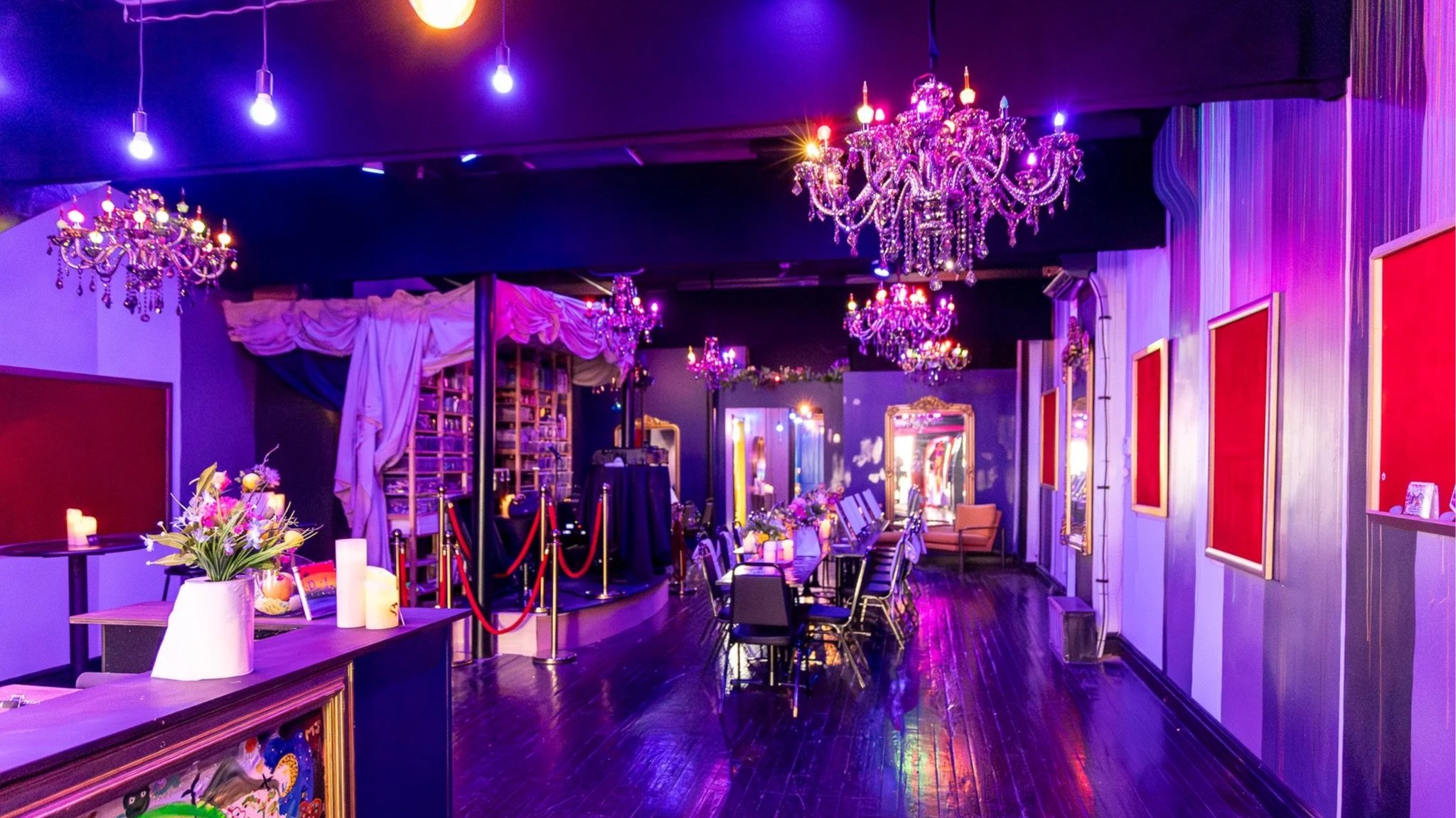 Interior of a decorated event space with crystal chandeliers, floral arrangements, and tables set for a celebration, illuminated with colorful purple and pink lighting.