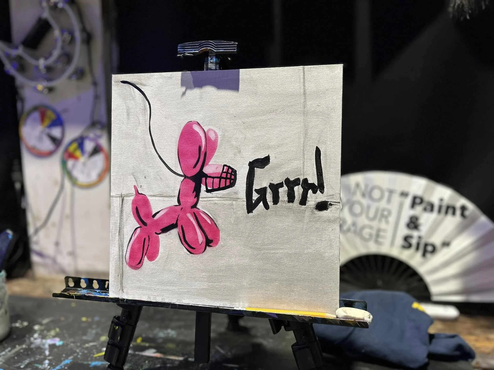 A small art piece on an easel featuring a pink cartoon-style dog dog with a black outline and minimal details, situated in an art studio with various supplies around.