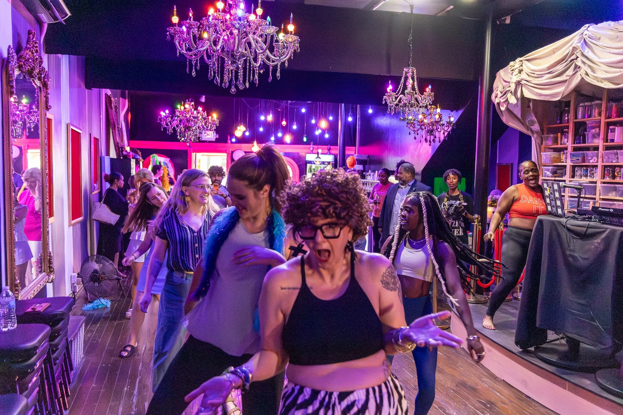 People dancing and socializing in a brightly lit nightclub with purple and pink lighting, chandeliers, and colorful hanging lights.