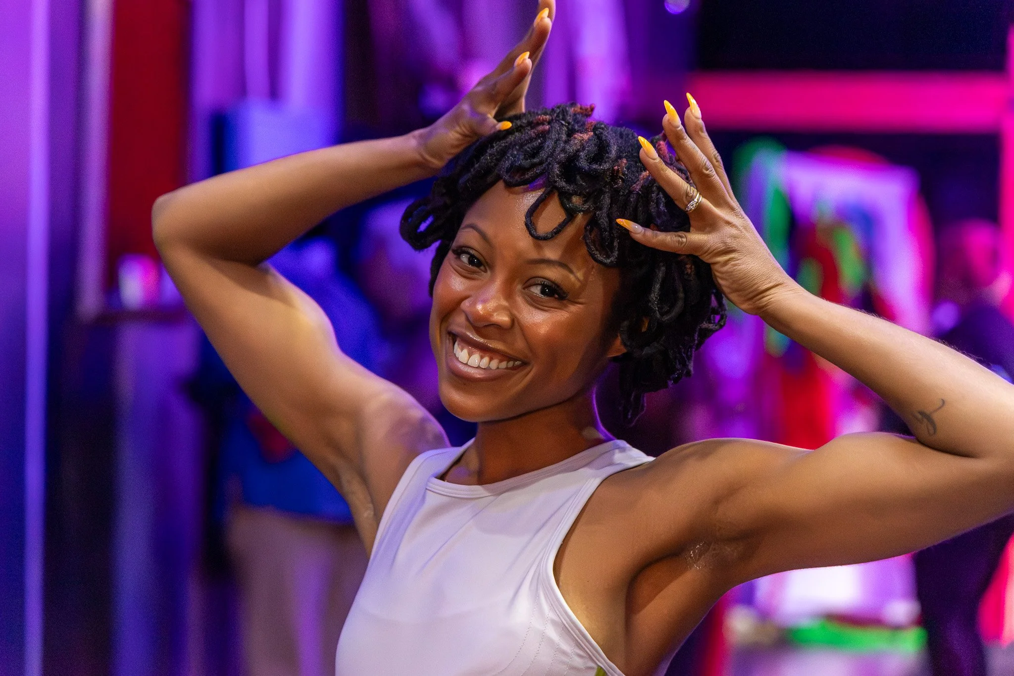 A woman smiling and posing with her hands on her head in a colorful, vibrant setting with purple and pink lighting.