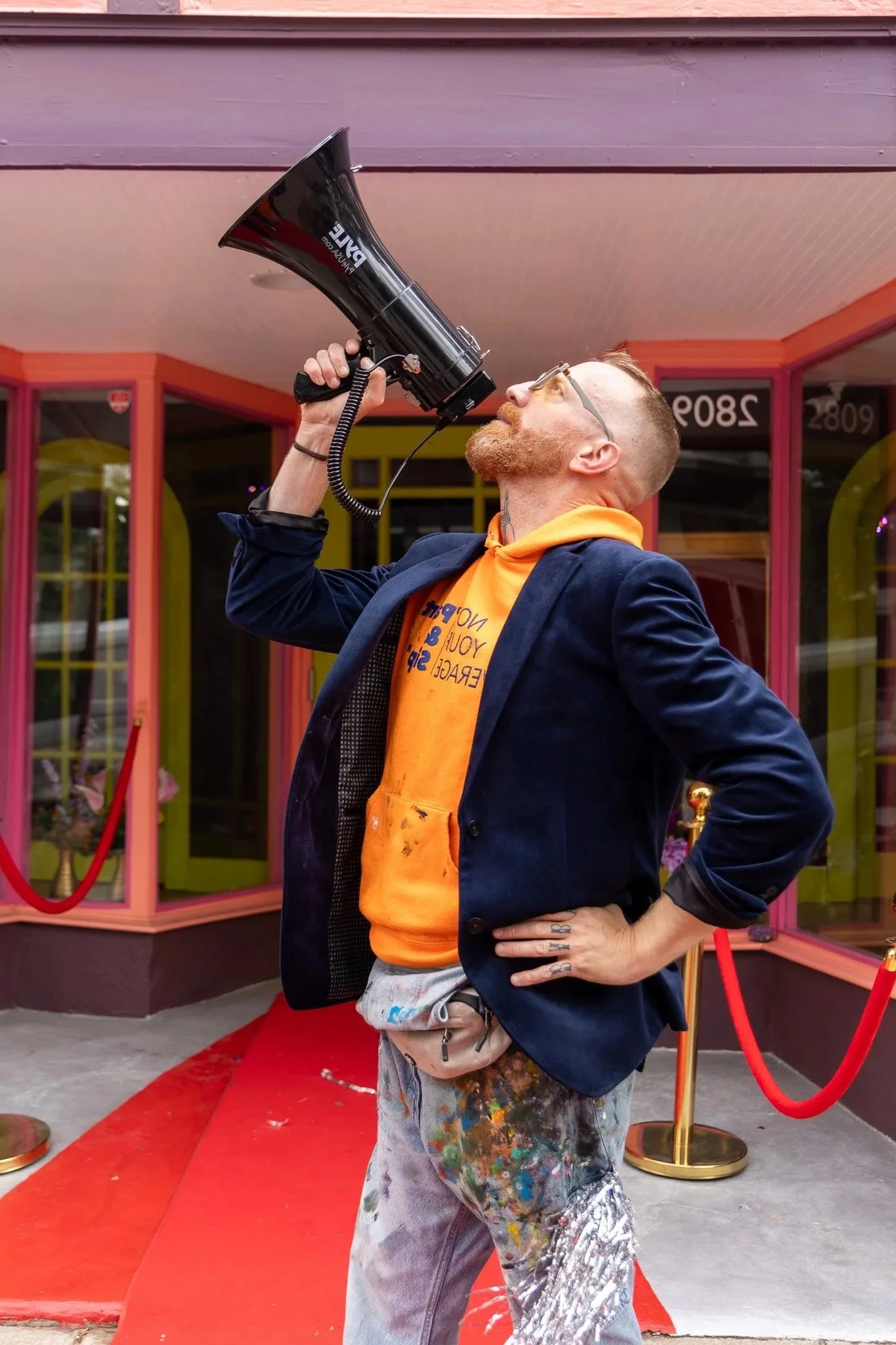 A man holding a large black megaphone to his mouth, standing on a red carpet outside a building with colorful glass windows, wearing glasses, an orange hoodie, a navy blazer, and paint-splattered pants.