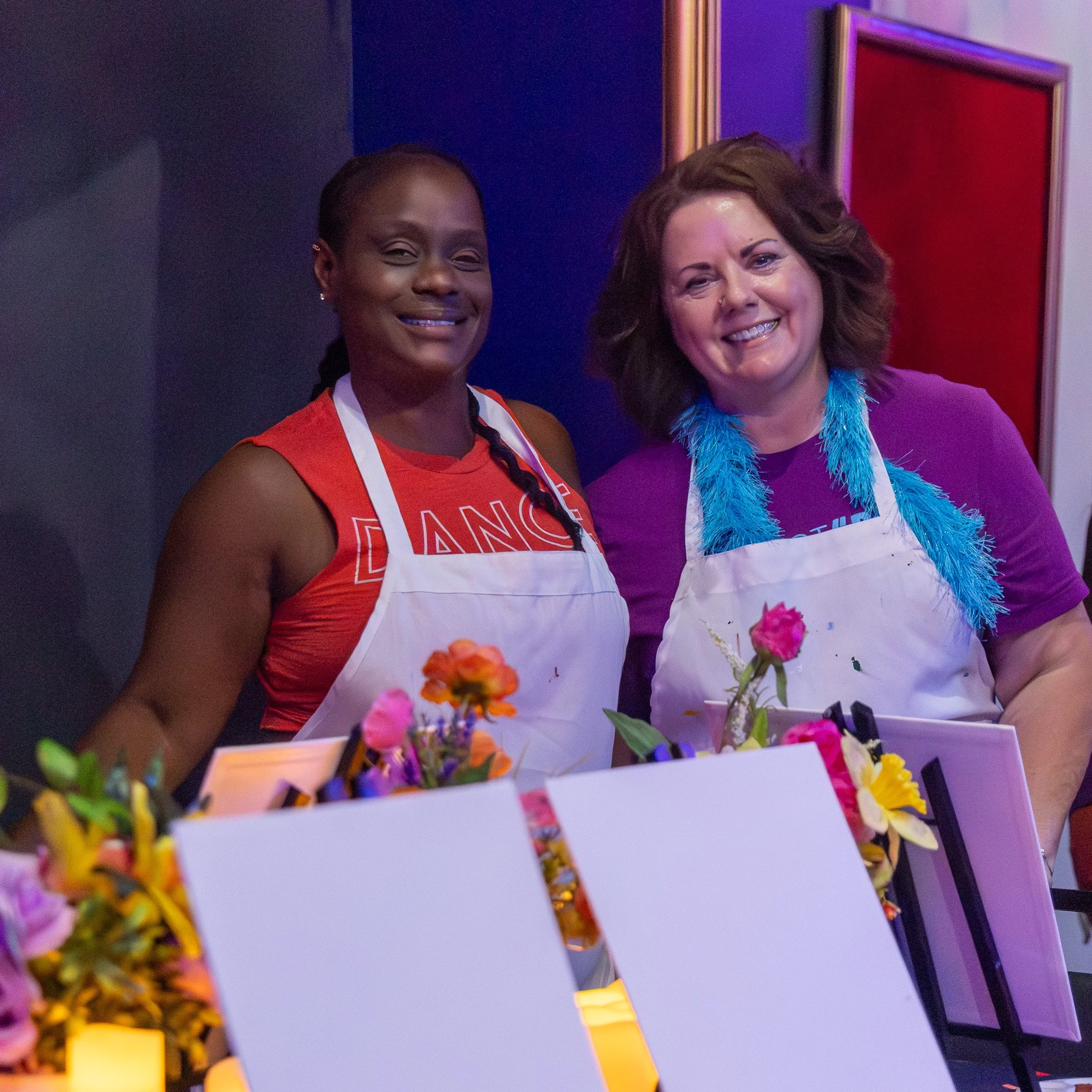 Two women smiling, standing behind a table with colorful flowers and a decorated cake in front of them. One woman has dark skin and wears a red shirt with an apron, while the other has lighter skin and wears a purple shirt with a fluffy blue scarf. The background features framed artwork on the wall.