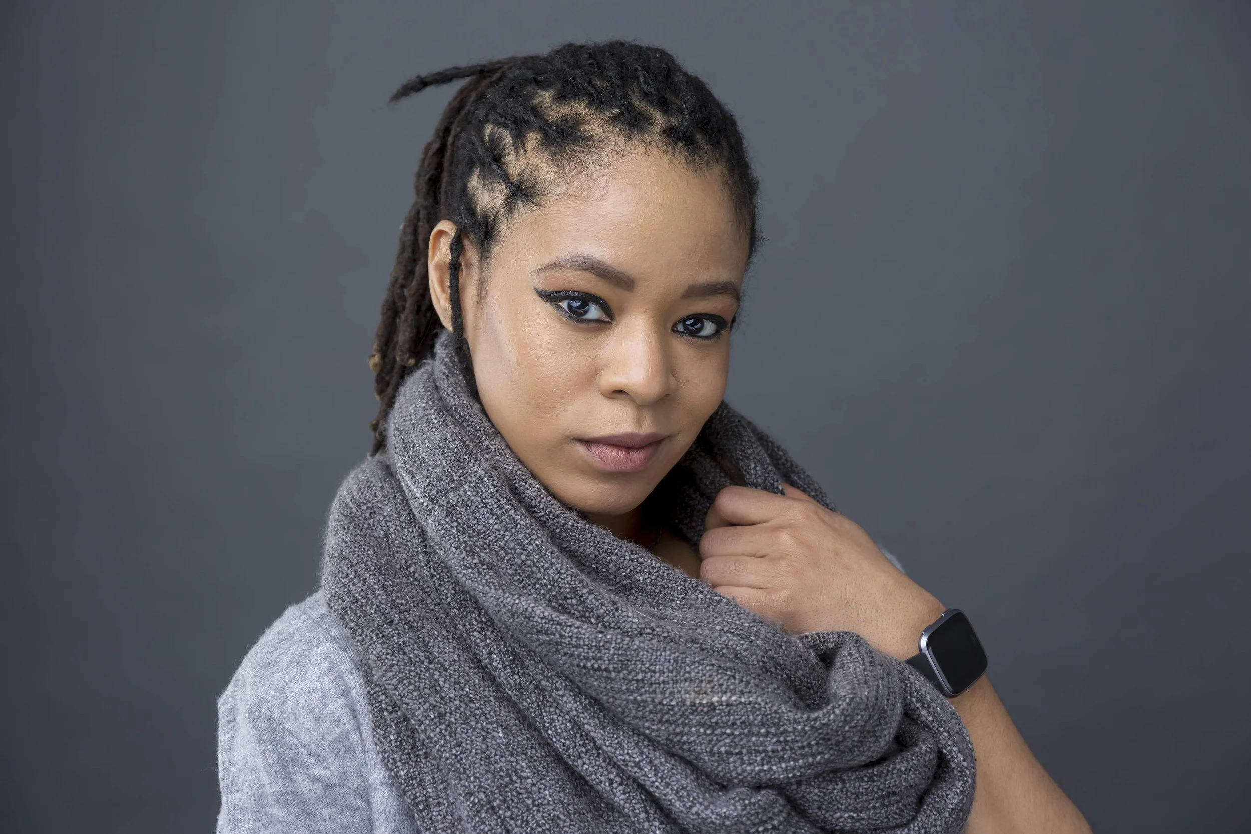A woman with dreadlocks wearing a gray scarf and smartwatch, posing against a gray background.