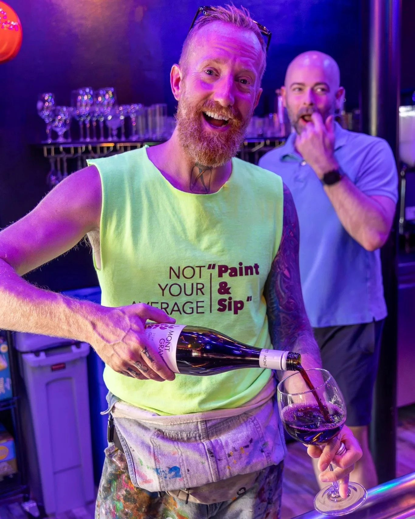 A man with a red beard and tattoos on his arm, wearing a yellow tank top that says "Not your average sip & paint," is pouring red wine into a glass. Behind him are another man with a bald head, wearing a gray shirt, and glasses on his head, who is holding a hand to his mouth. The scene is lit with purple and blue lighting, and glasses and bottles are visible in the background.