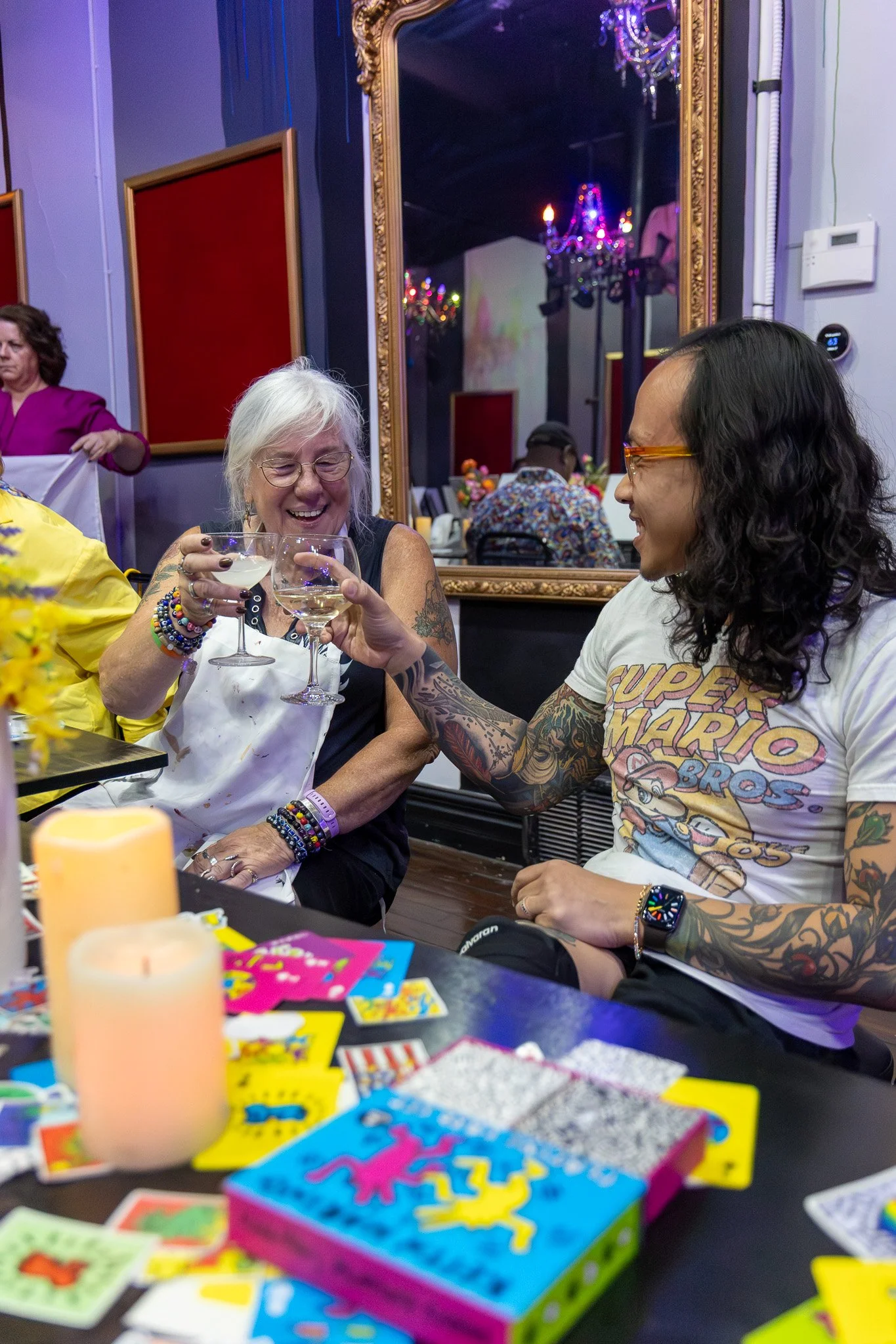 Two women smiling and clinking glasses of white wine at a party or gathering, with a table full of colorful board games and a large ornate mirror in the background.