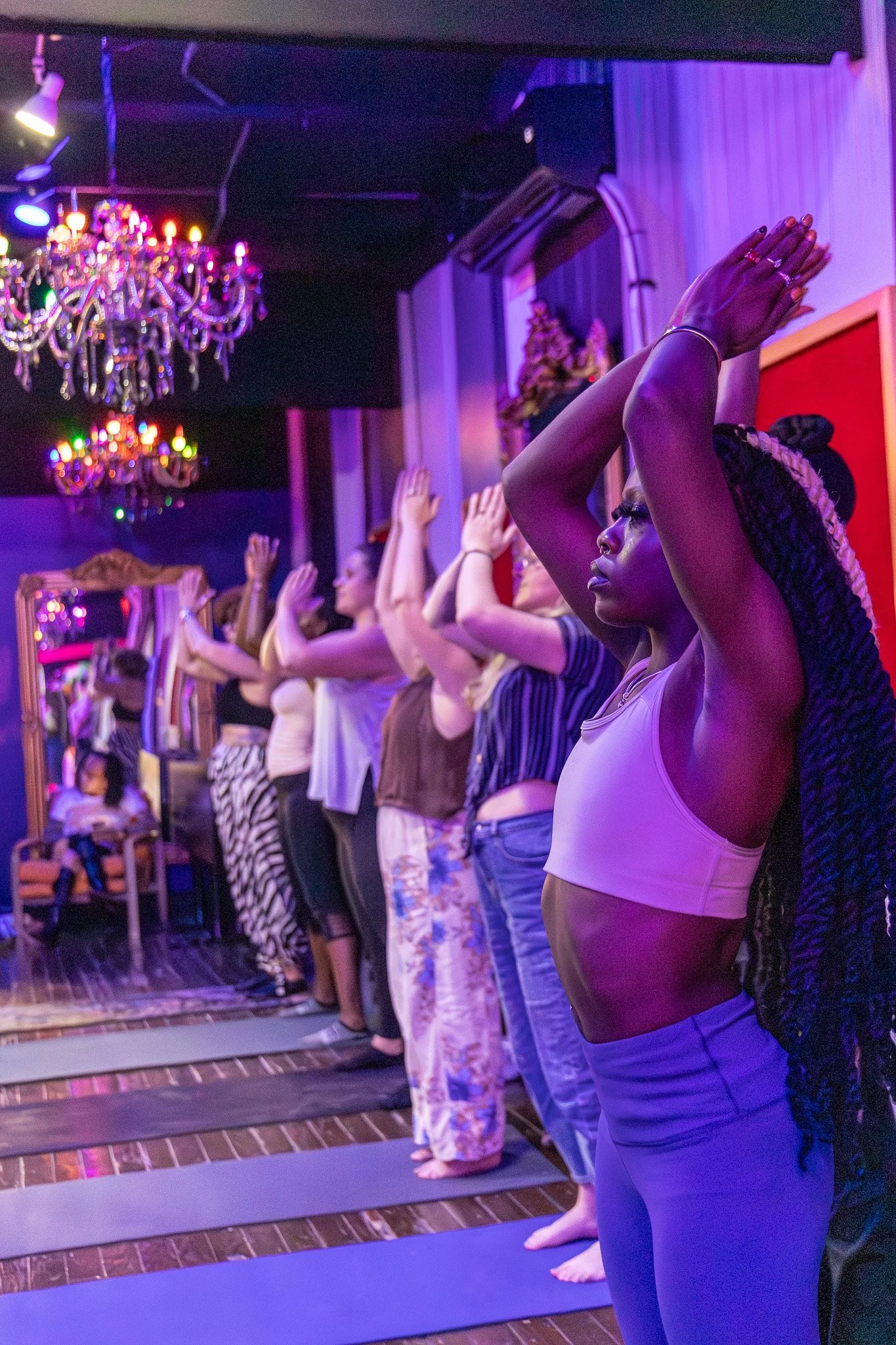 A diverse group of people practicing yoga in a dimly lit room with purple and pink lighting, standing on yoga mats with hands raised overhead, some with closed eyes, in a peaceful setting with chandeliers and decorative elements.