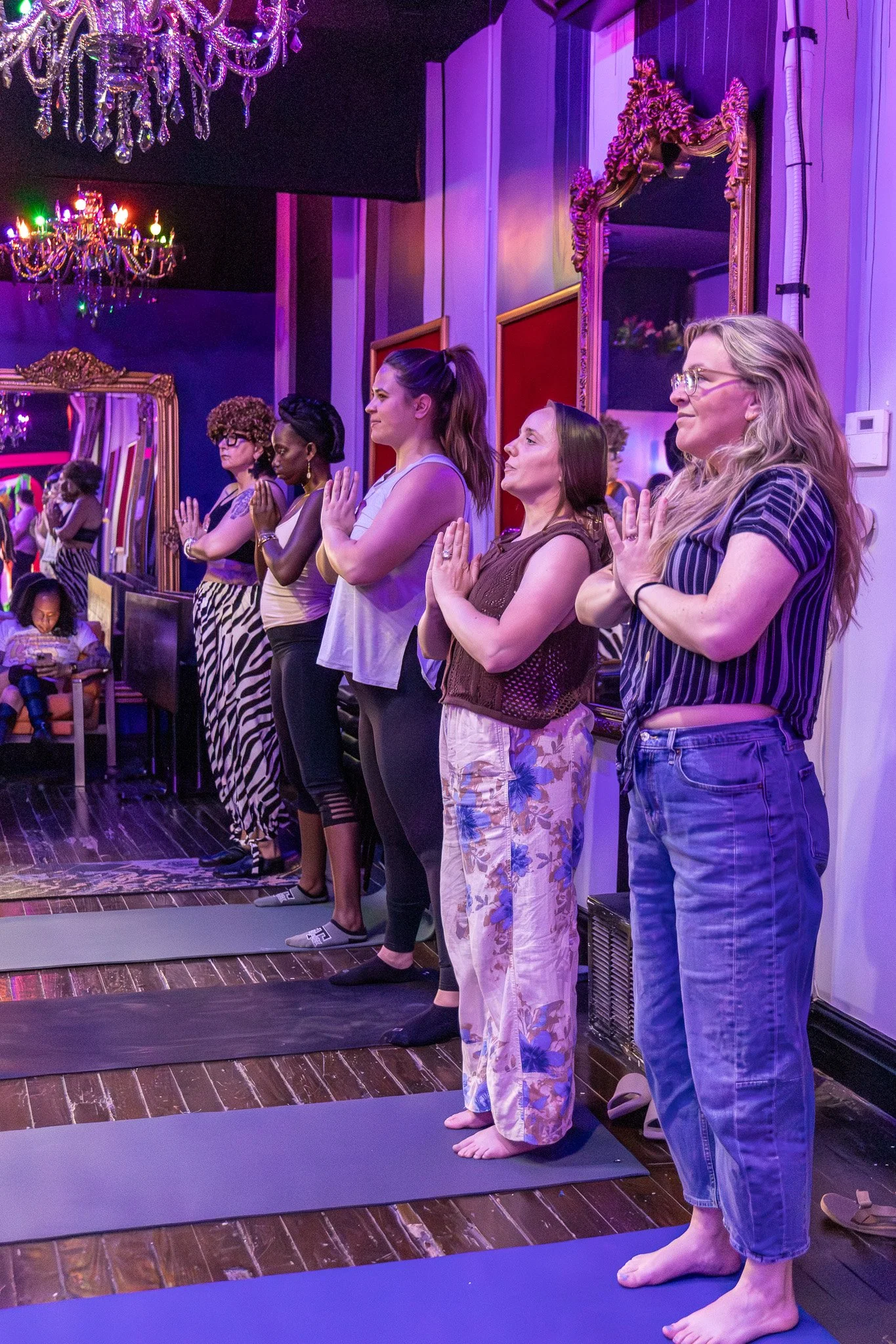 A group of five women practicing yoga or meditation together in a room with purple lighting and chandeliers. They are standing on mats with their hands in prayer position and are facing forward. The women are dressed in casual, comfortable clothing, some barefoot, in a peaceful and focused manner.