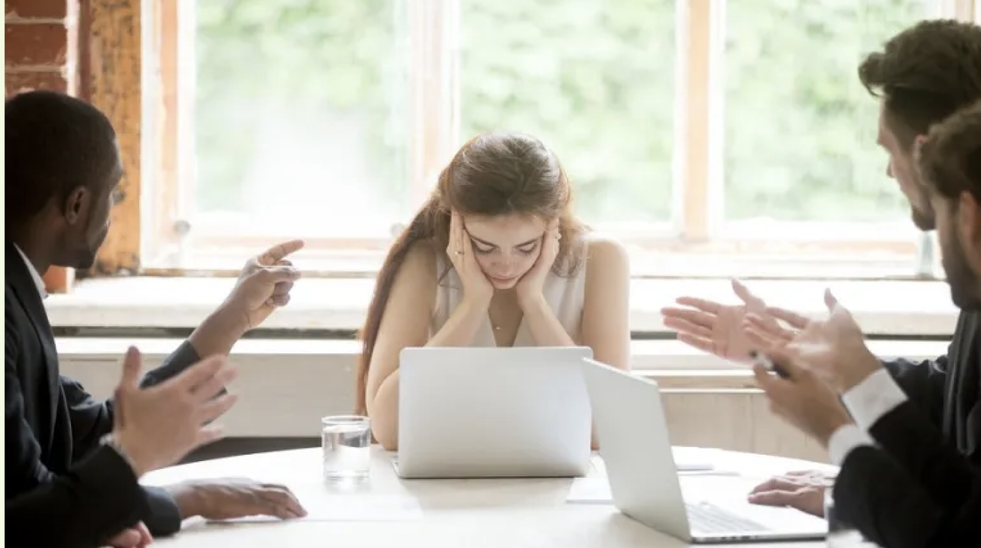 Young woman frustrated with her head in hands sitting at a table with a laptop, while three men in suits on either side are gesturing and appear to be arguing or debating with her.