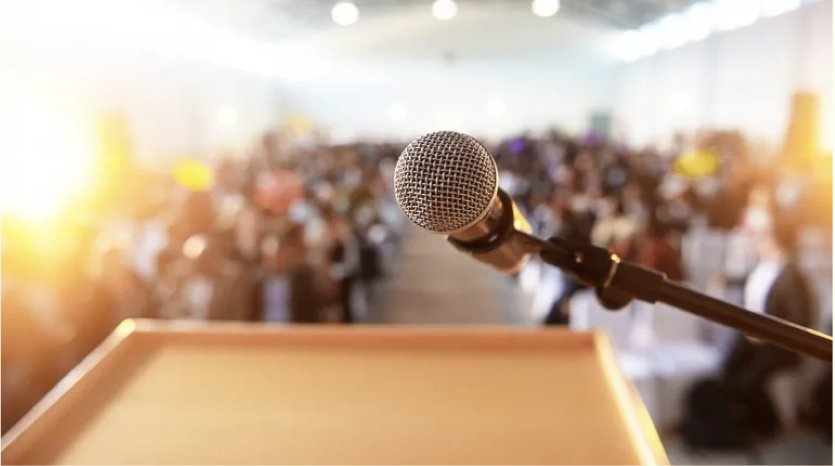 A close-up of a microphone on a stand in front of a large audience in an auditorium or conference room, with sunlight streaming in from the left side.