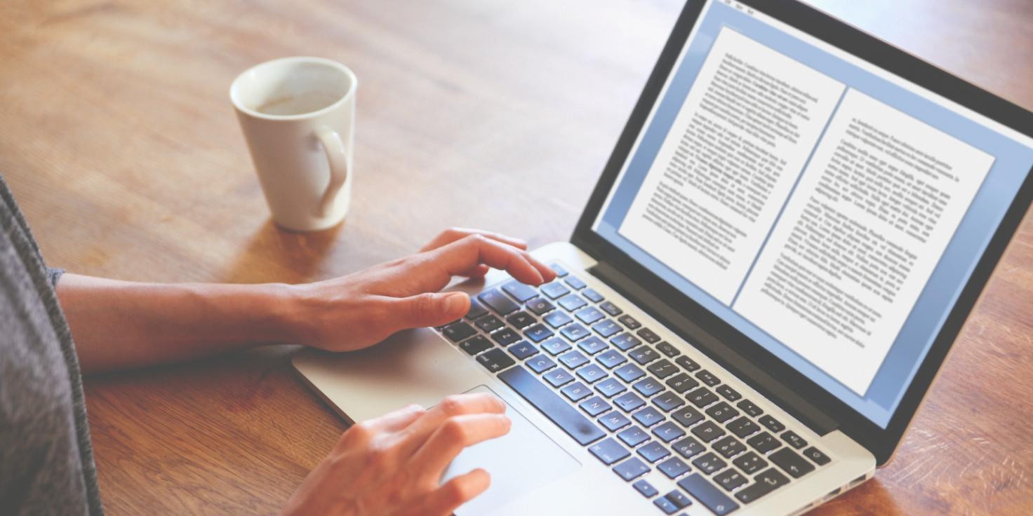 Person typing on a laptop with an open document on the screen, a white mug nearby, and a wooden table surface.