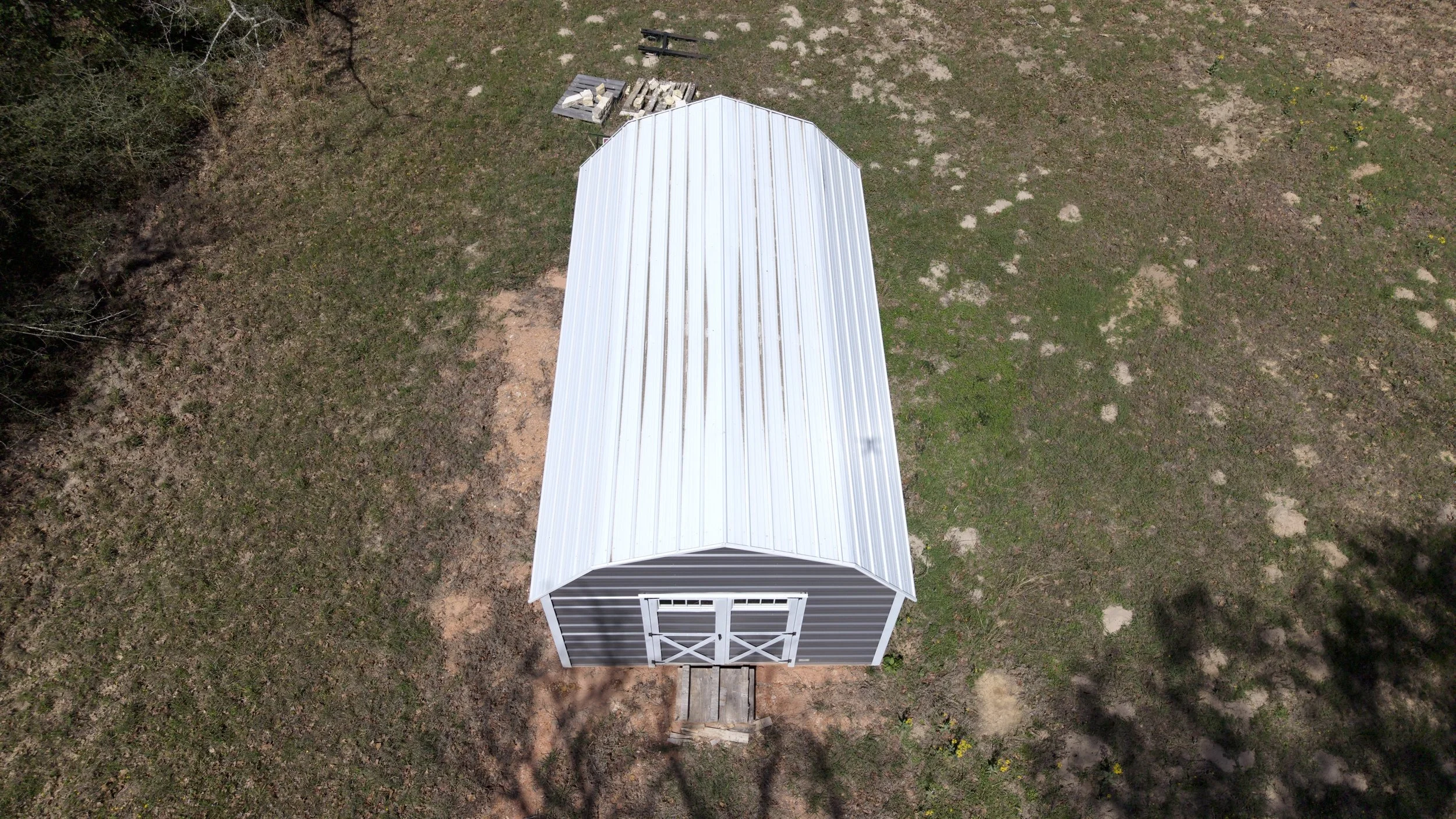 A small gray and white shed with a metal roof, viewed from above on a grassy area with patches of dirt.