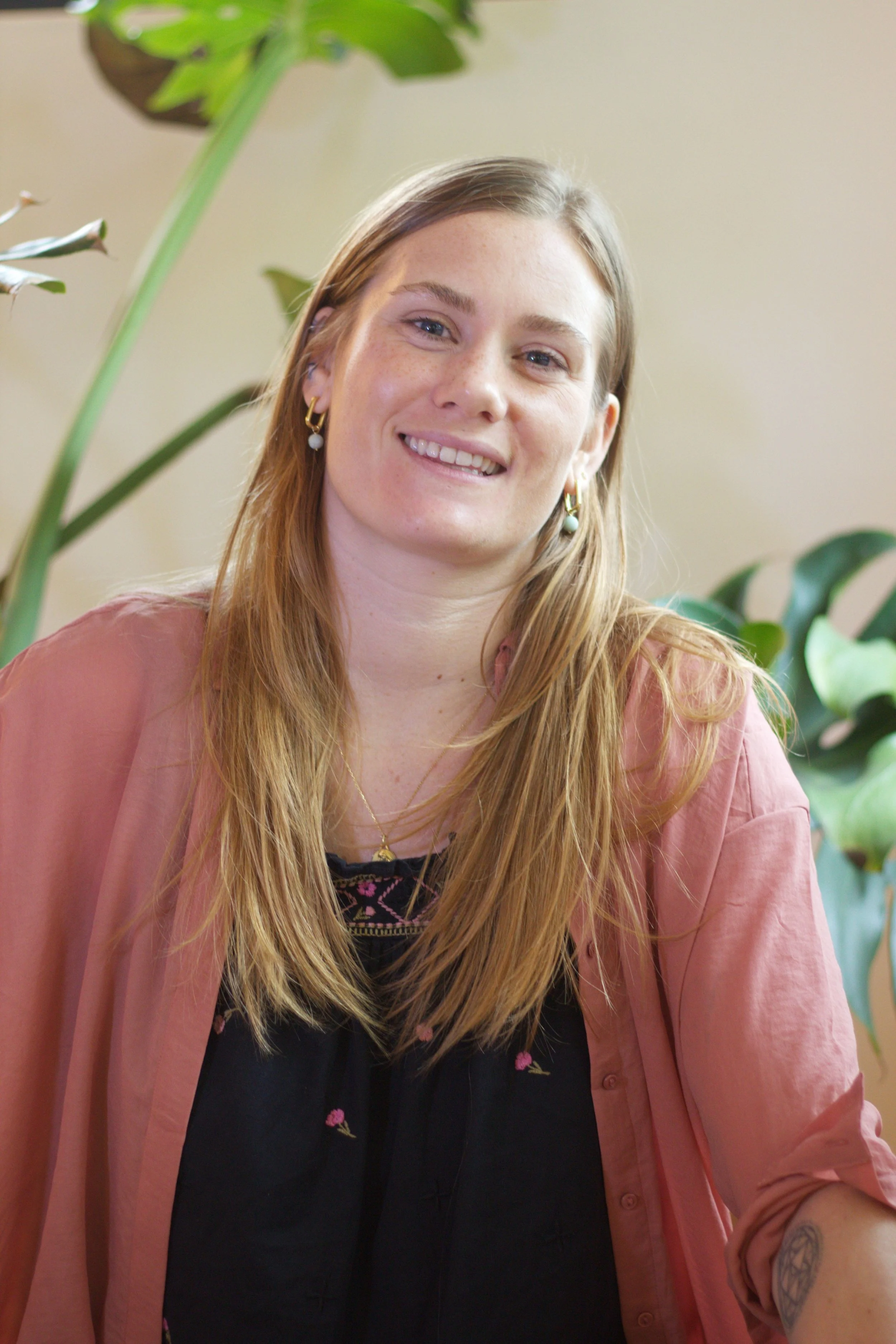 A young woman with long reddish-blonde hair smiling, wearing earrings, a necklace, a black top with pink embroidery, and a pink jacket, sitting near large green houseplants.