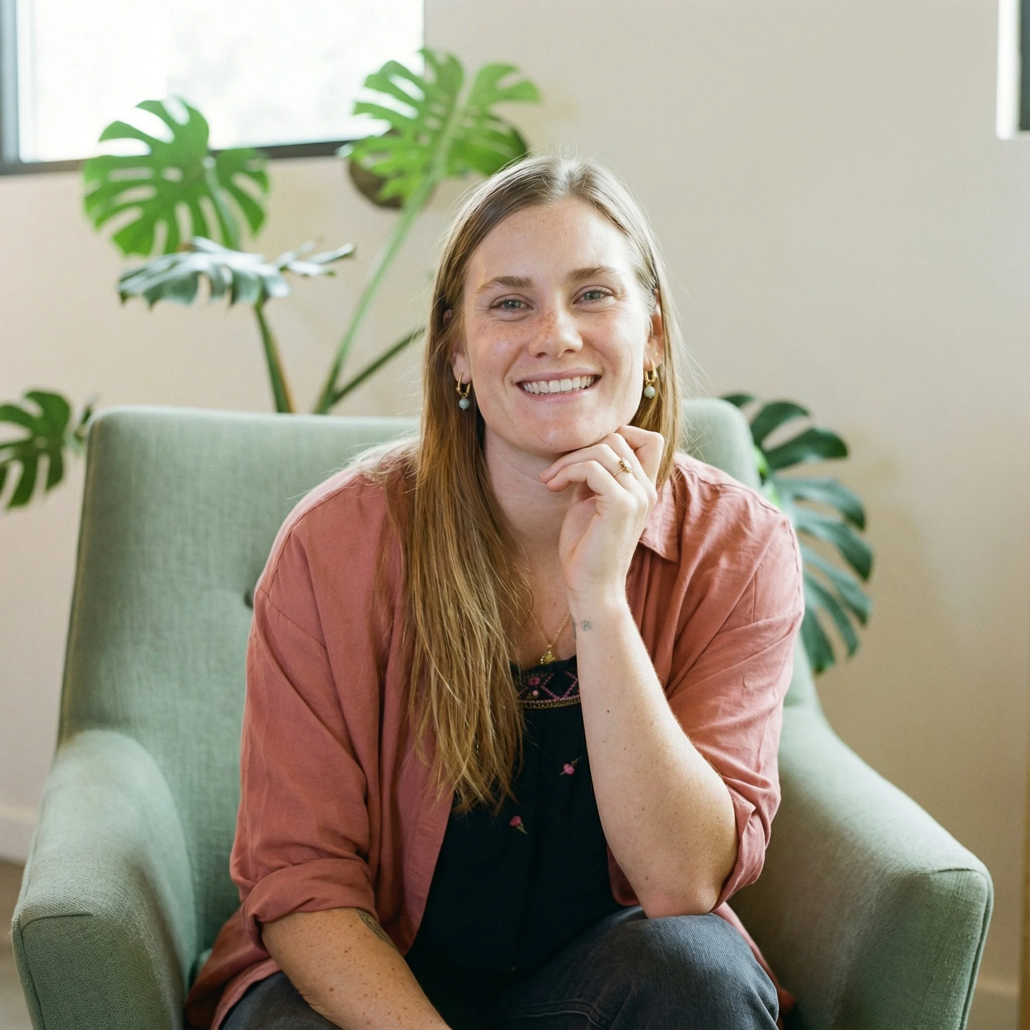 A woman with long blond hair, freckles, and earrings sitting in a green armchair, smiling with her chin resting on her hand, in a room with a large monstera plant behind her and a window.