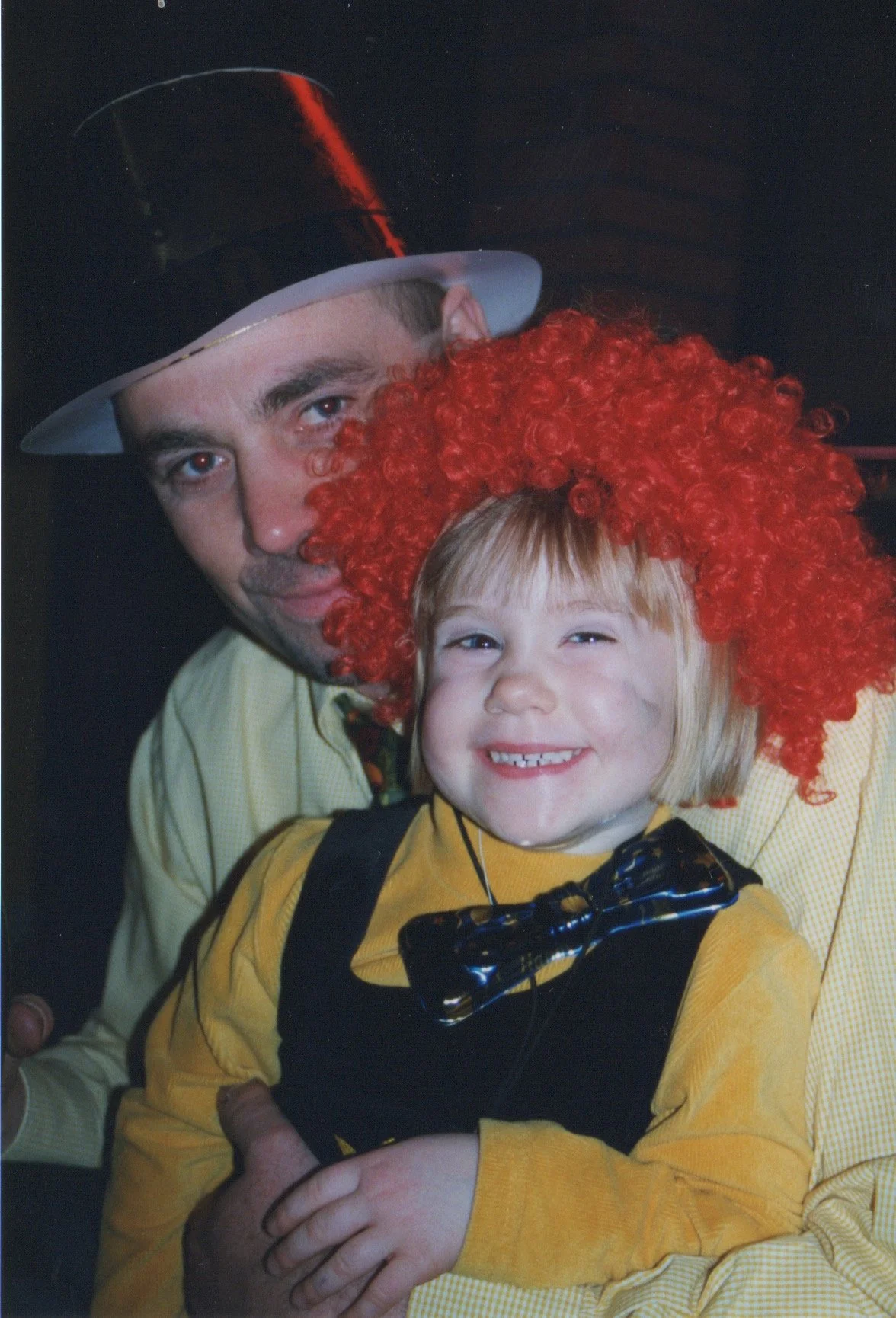 A man wearing a hat with a red feather and a young girl with a curly red wig and a yellow shirt with a black bow tie, smiling and hugging each other.